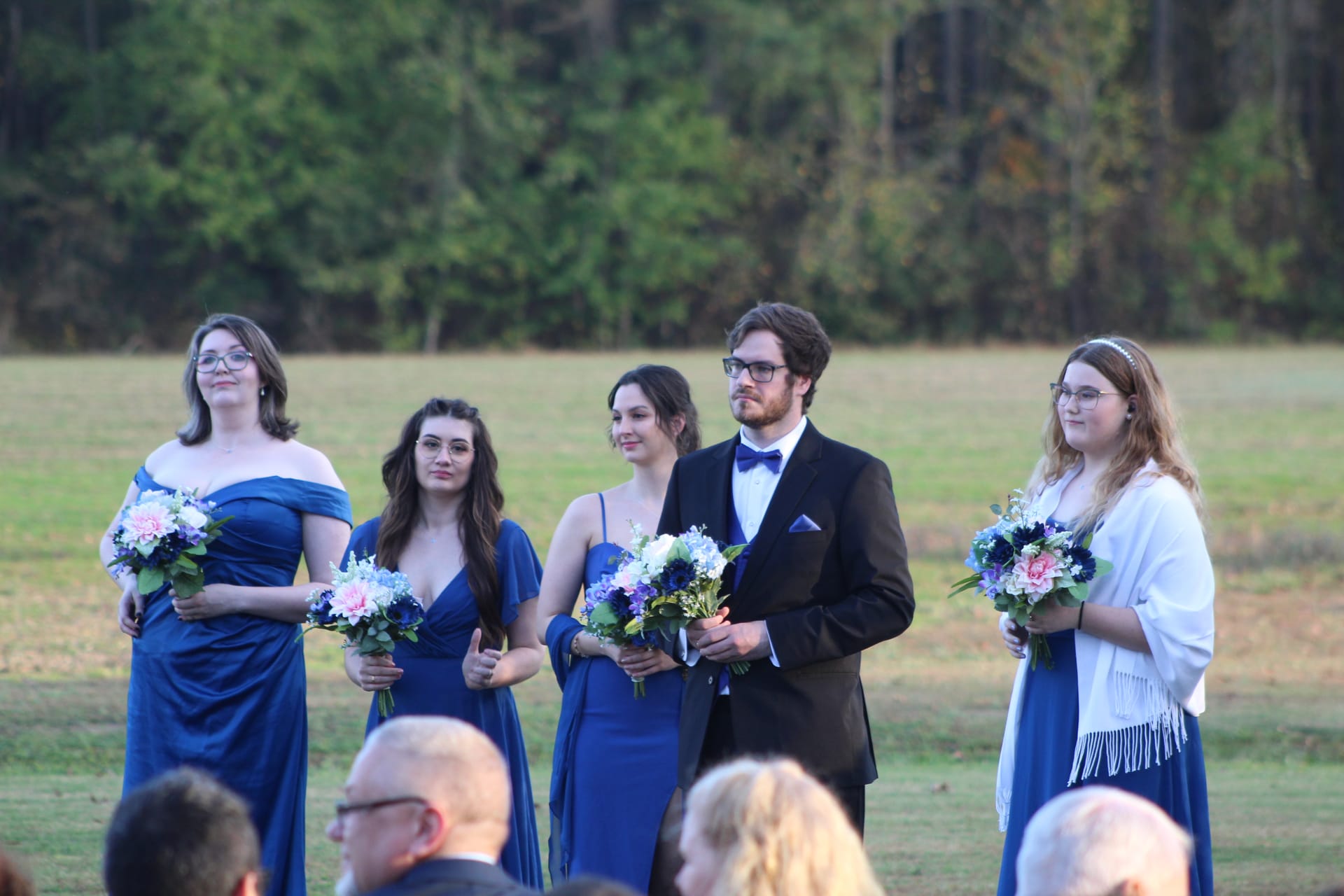 Three bridesmaids in blue gowns and a groomsman in a black tuxedo hold floral bouquets while standing on the ceremony lawn at Scarborough House, with a wooded backdrop behind them.