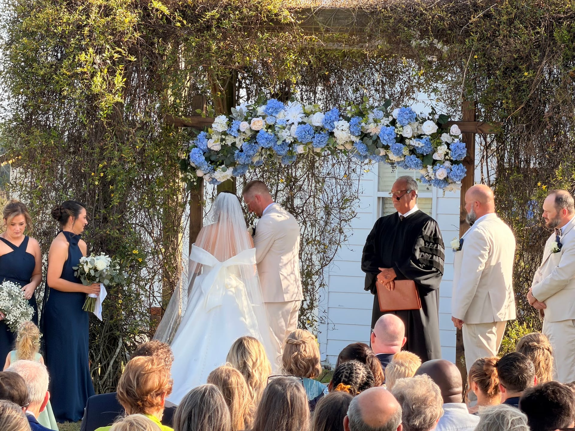 Bride and groom share an intimate moment beneath a lush floral arch adorned with blue hydrangeas and white roses during an outdoor wedding ceremony at Scarborough House, surrounded by bridal party and seated guests.