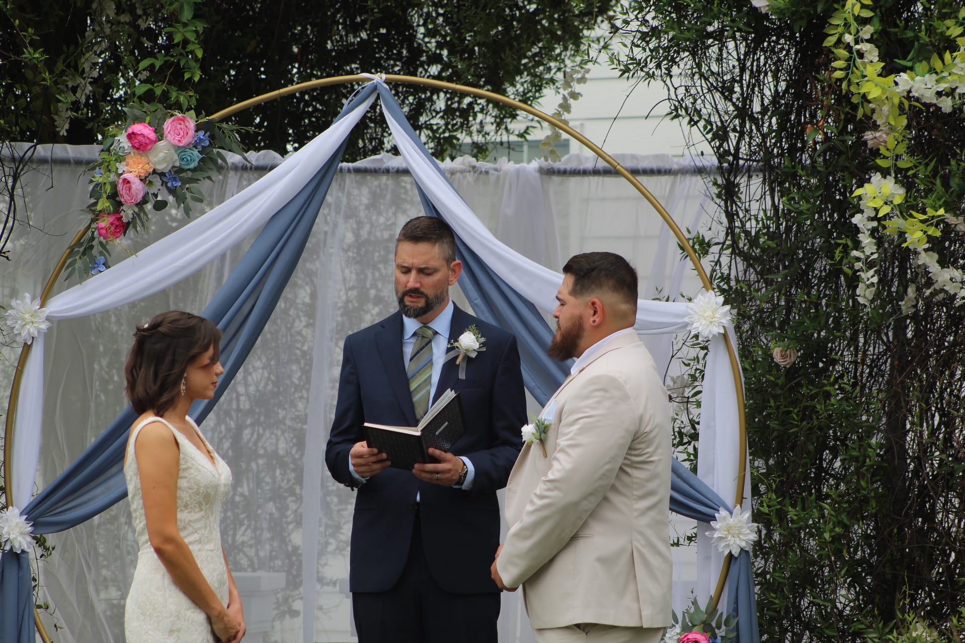 Bride and groom stand before an officiant reading from a book beneath a gold circular arch adorned with blue draping and colorful florals during an outdoor wedding ceremony.