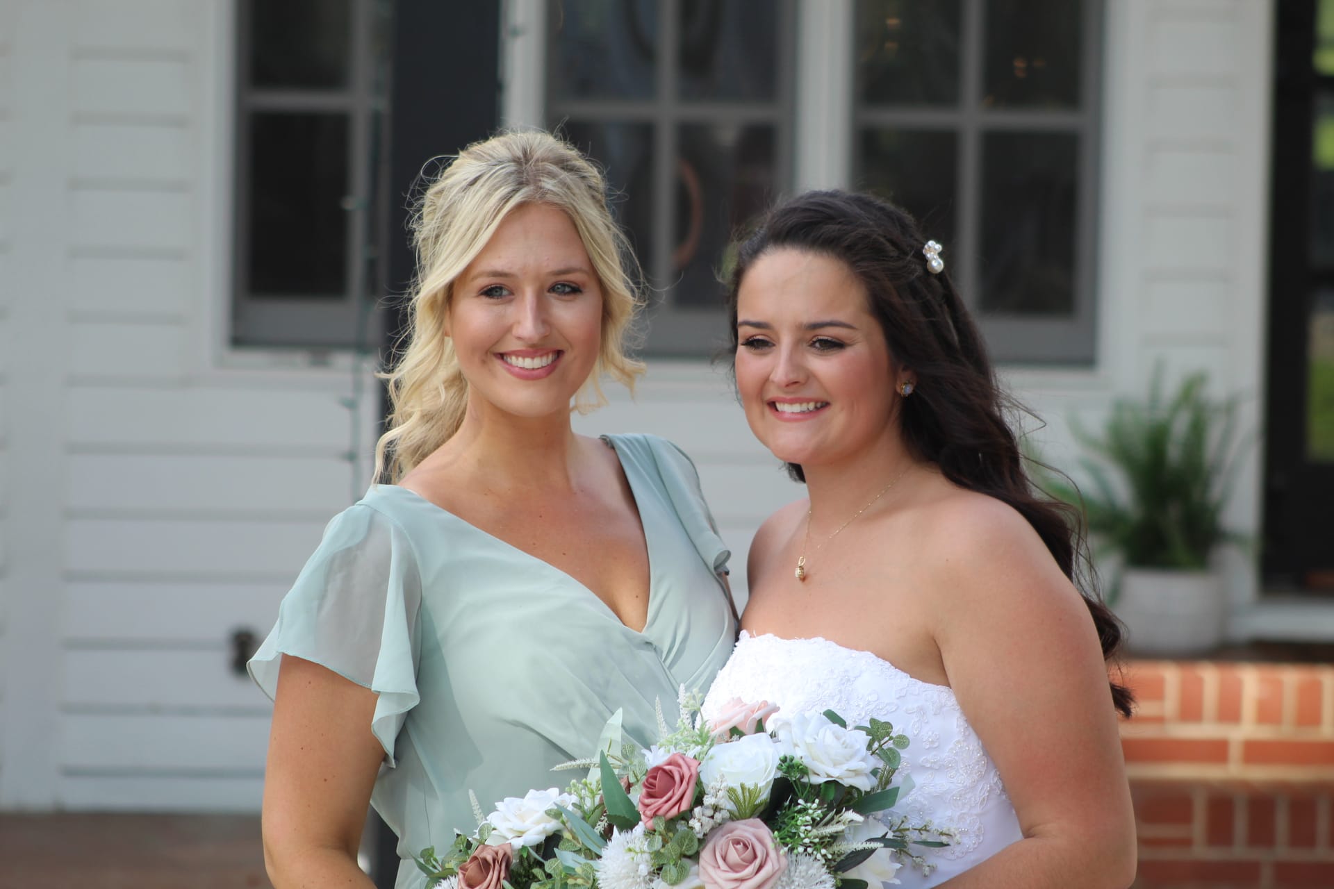 Bride in a strapless white gown holding a bouquet of blush and white roses stands with a bridesmaid in a sage green dress in front of the historic white manor house at Scarborough House.