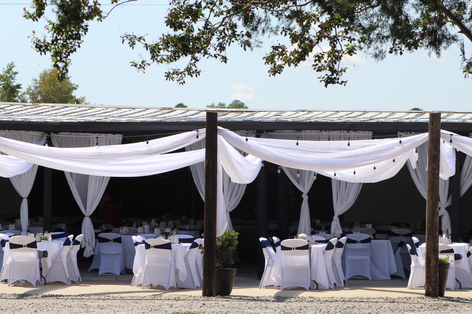 Elegantly decorated outdoor wedding reception pavilion at Scarborough House featuring flowing white fabric draping, string lights, white chair covers with navy sashes, and round guest tables set for a celebration.
