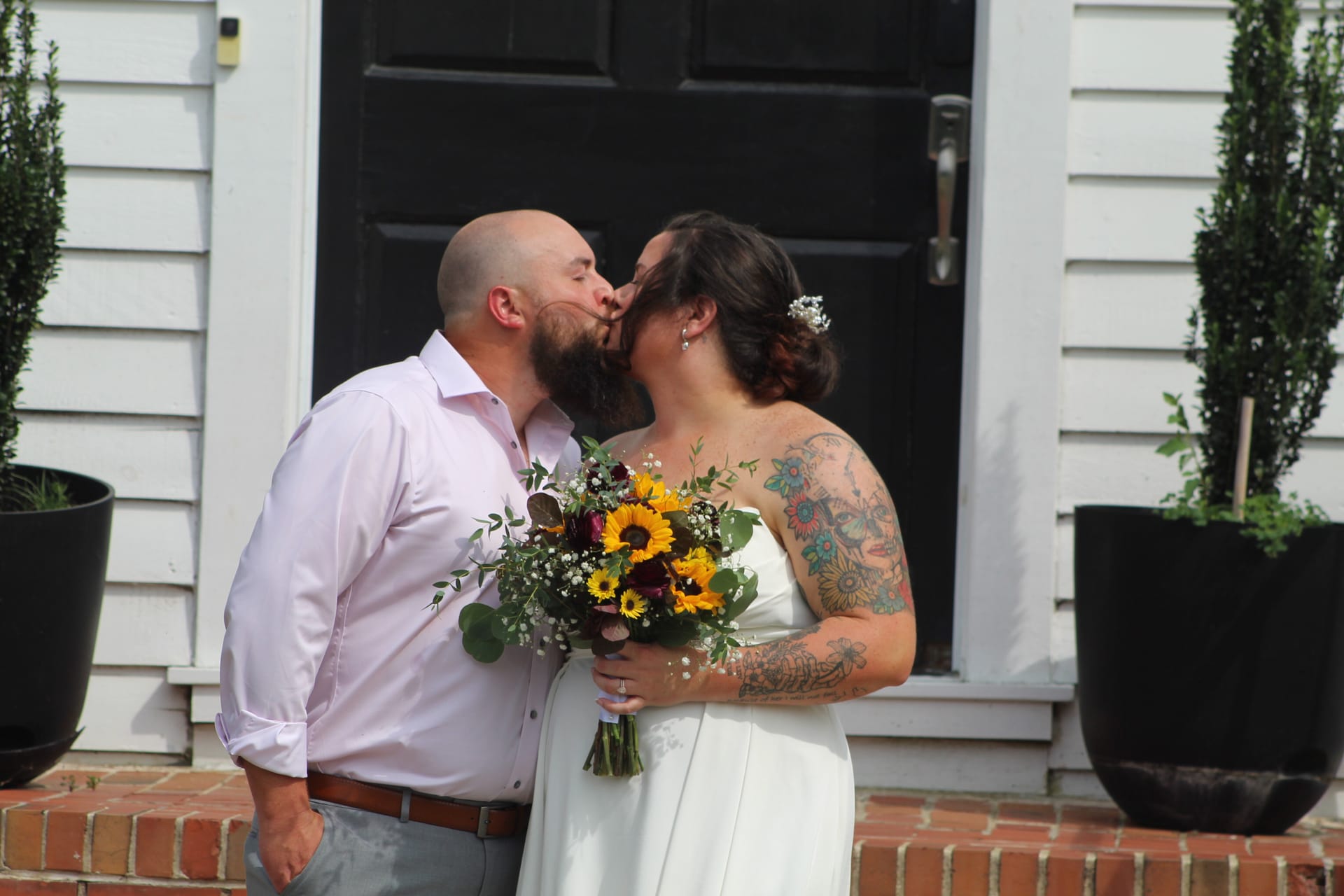 Newlyweds share a first kiss on the brick front steps of the 1821 Scarborough House manor, the bride holding a vibrant sunflower and greenery bouquet while wearing a white strapless gown.