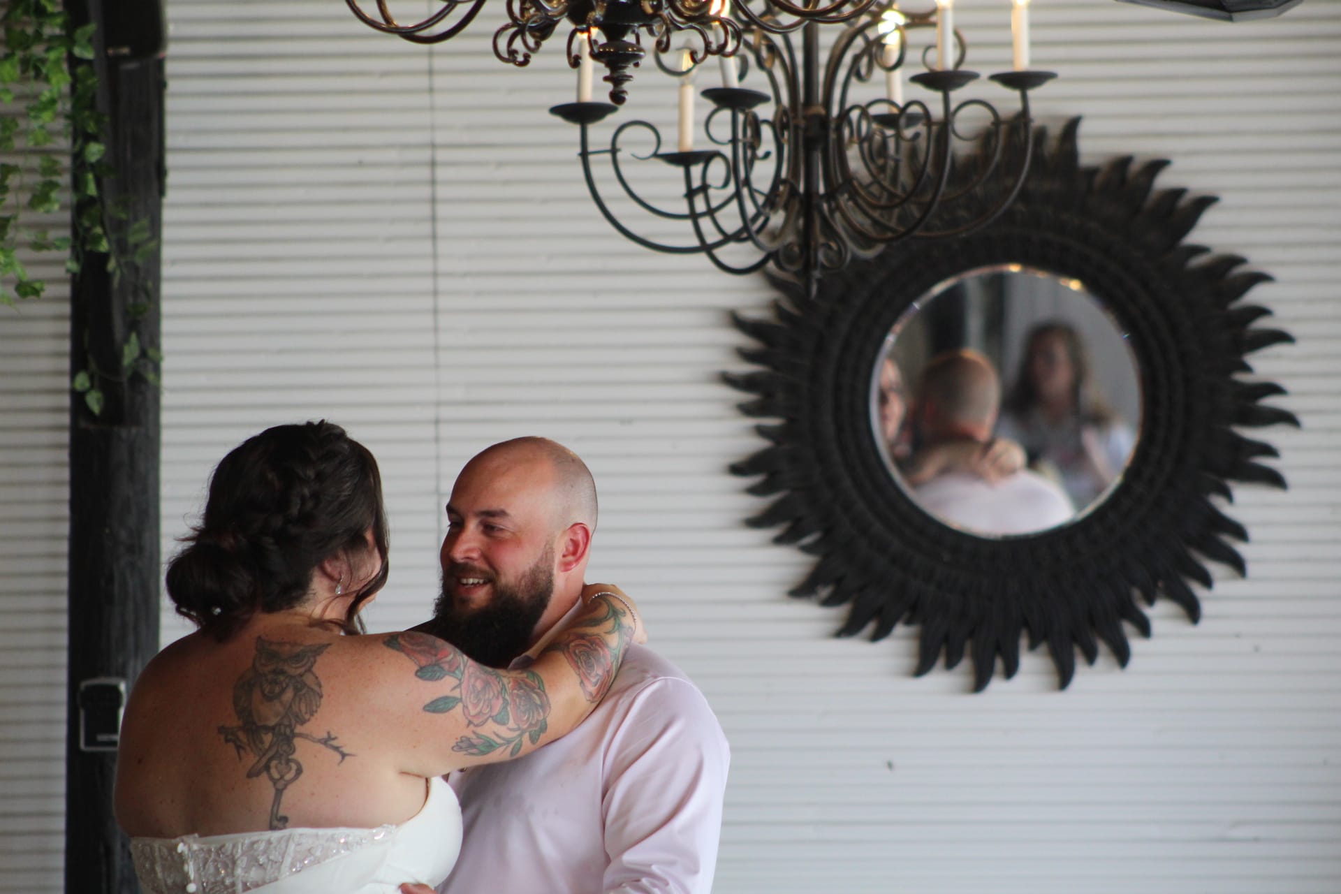 Bride and groom sharing their first dance beneath a wrought iron chandelier at Scarborough House, their embrace reflected in a decorative sunburst mirror on the white shiplap wall behind them.