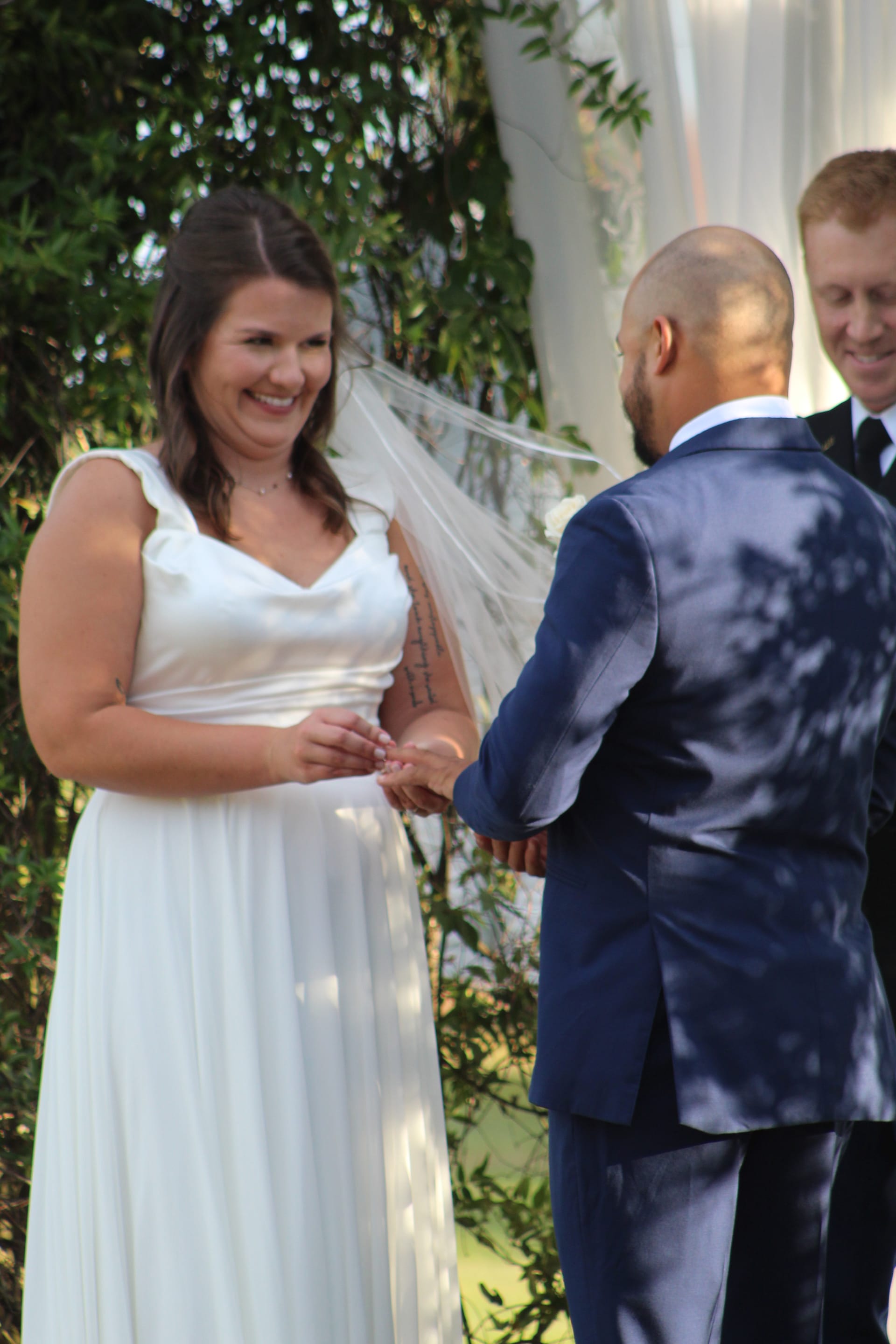 Smiling bride in a white gown and veil exchanges rings with her groom in a navy suit during an outdoor wedding ceremony at Scarborough House, surrounded by lush green foliage.
