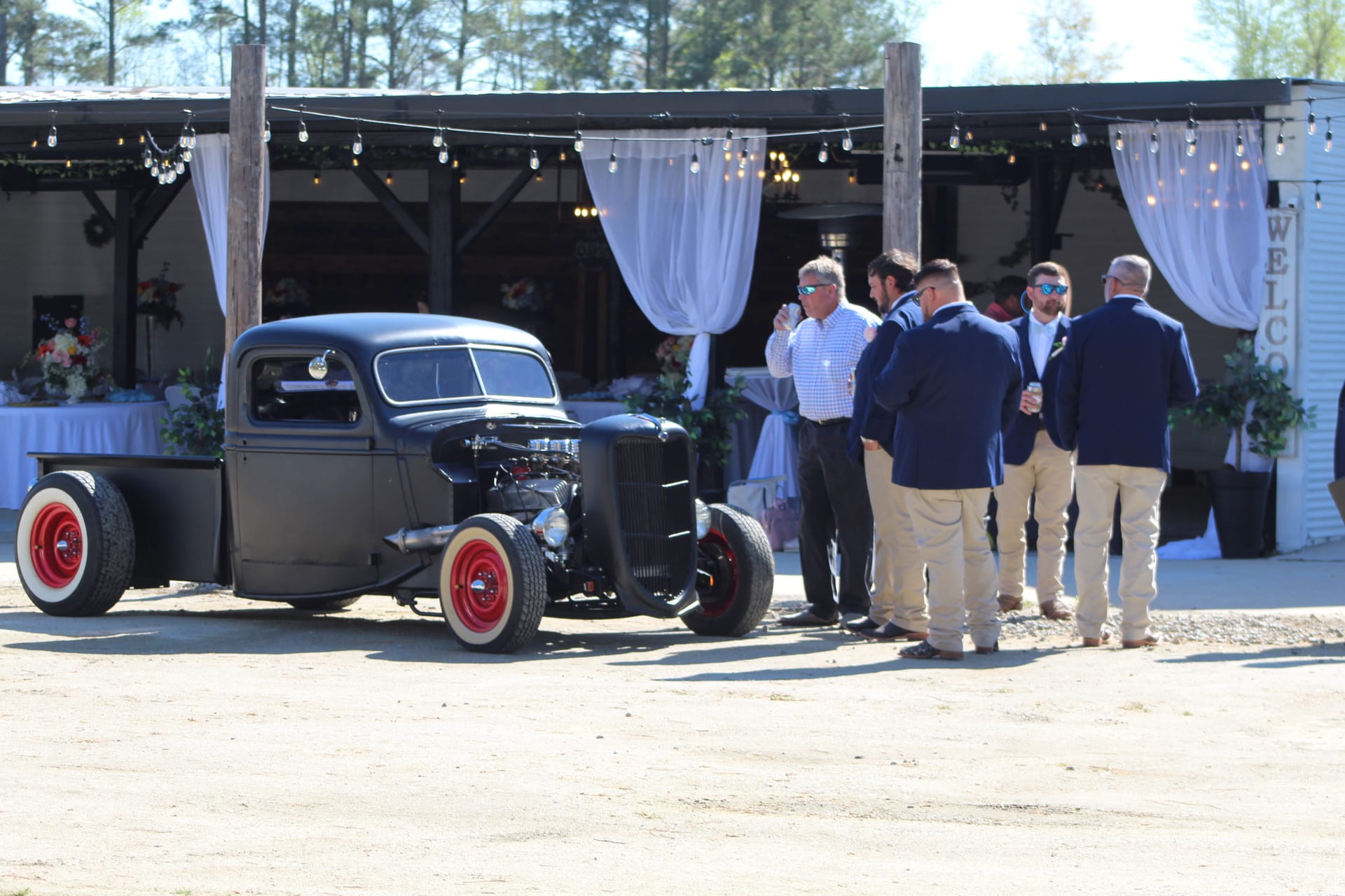 Groomsmen in navy blazers and khakis gather and chat beside a custom matte-black vintage hot rod truck outside a decorated open-air pavilion at Scarborough House wedding venue.