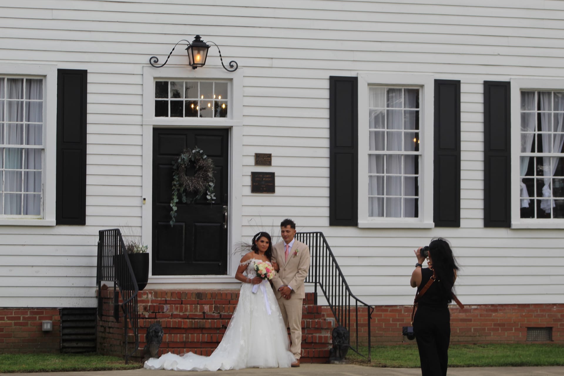 Bride in a white ball gown holding a pink bouquet and groom in a tan suit pose together on the front steps of the 1821 Scarborough House manor while a photographer captures their portrait.