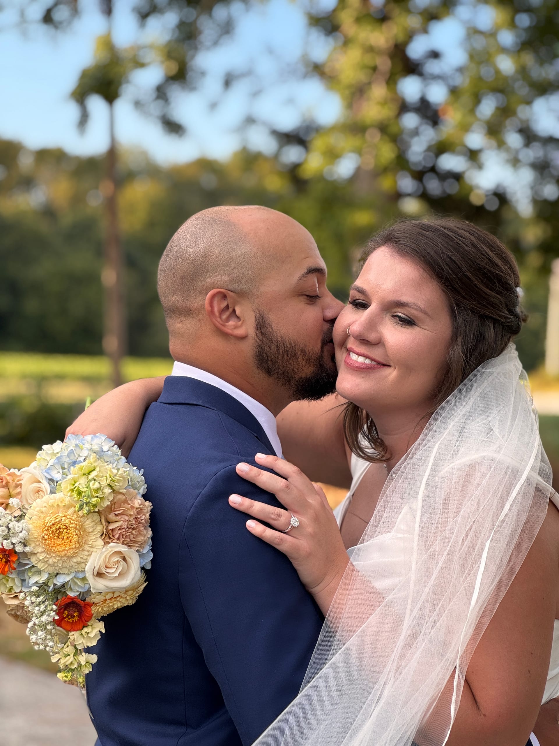 Groom in a navy suit kisses his beaming bride on the cheek as she holds a colorful bouquet of hydrangeas and garden roses beneath the oak trees at Scarborough House estate.