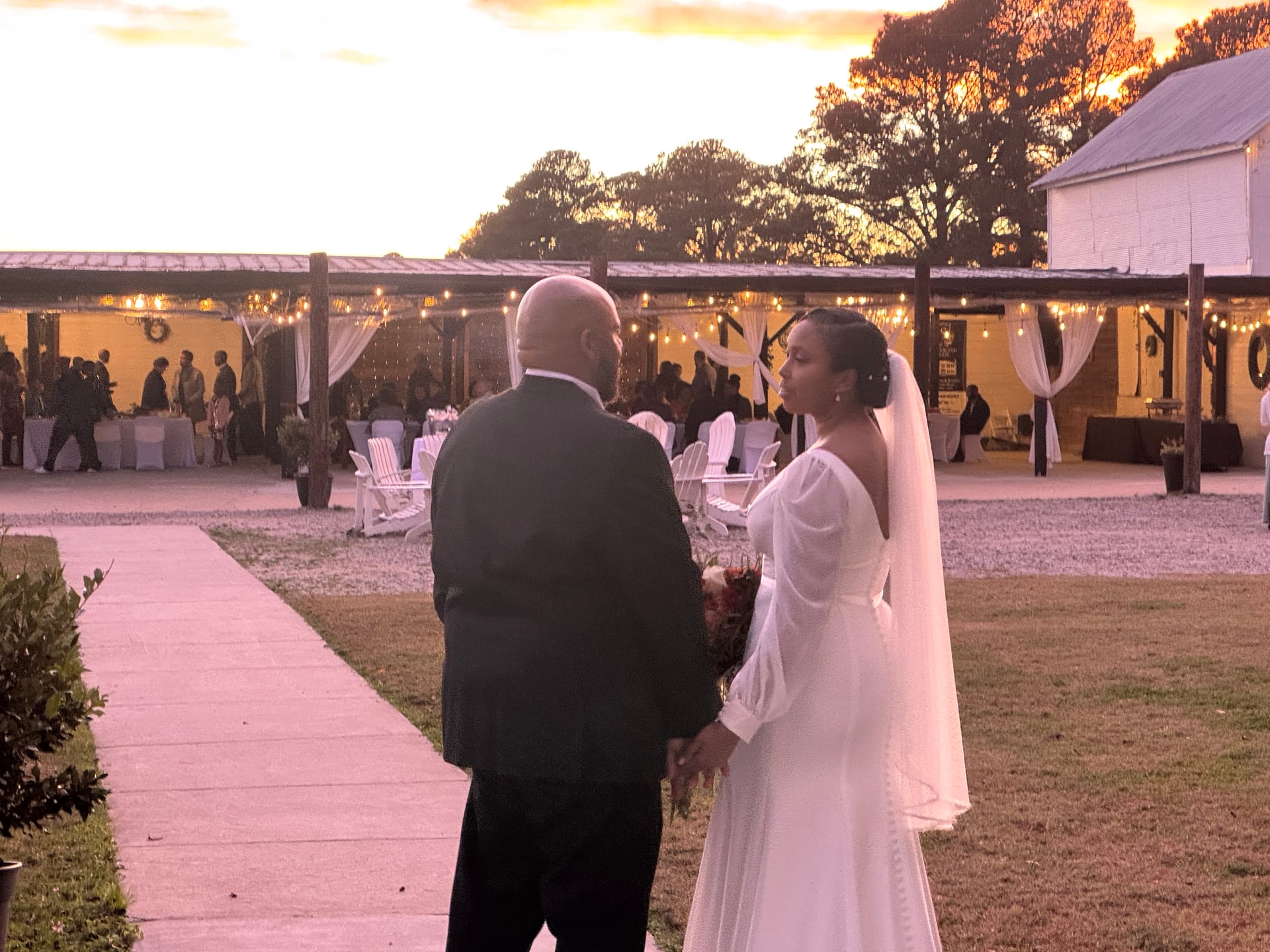 Bride in a flowing white off-shoulder gown with veil glances back while holding hands with her groom against a glowing golden-hour sunset at Scarborough House outdoor ceremony lawn adorned with string lights.