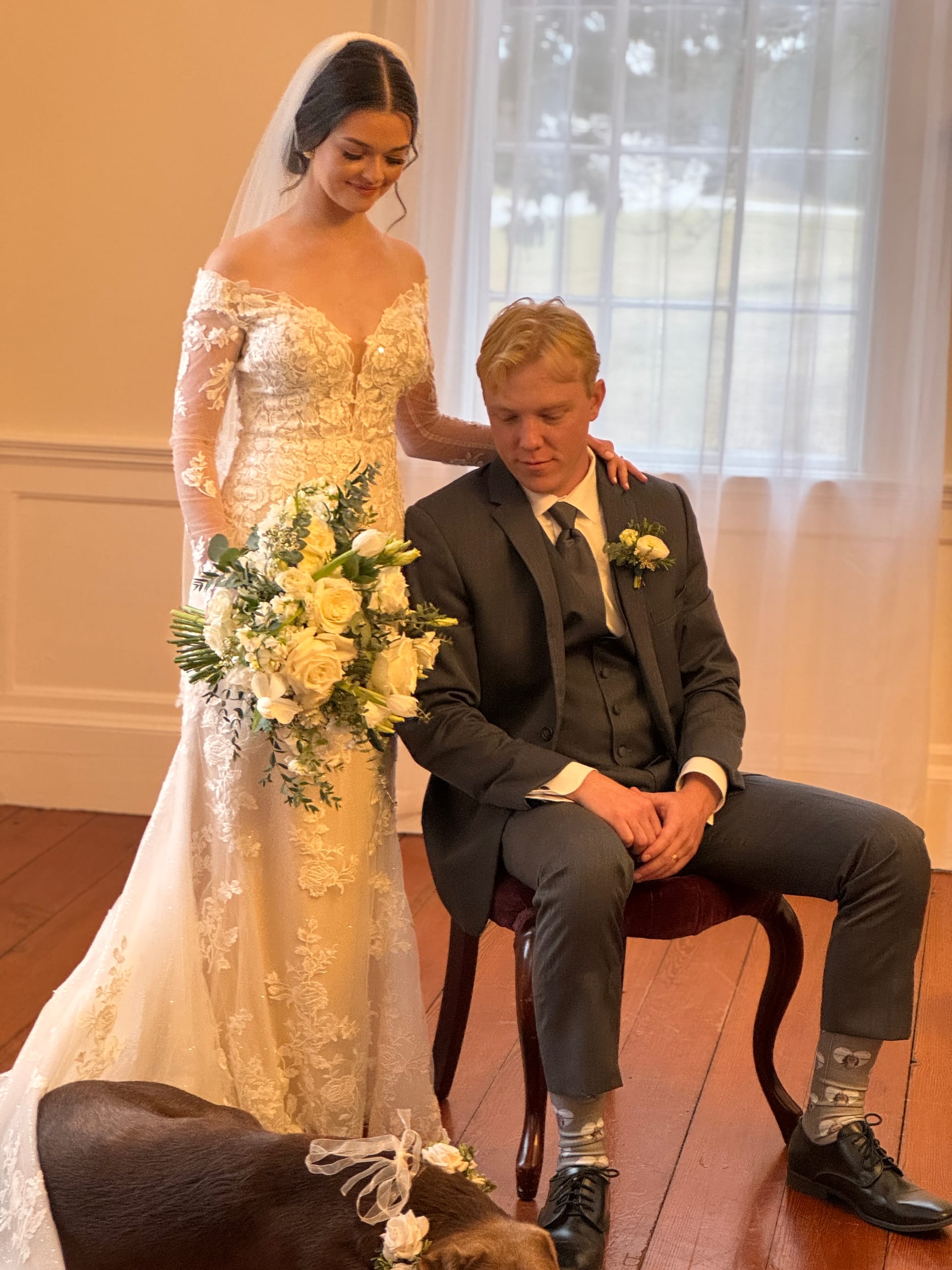 Bride in a lace off-shoulder gown holding a white and yellow floral bouquet stands behind her seated groom in a charcoal suit inside the historic Scarborough House grand hall.