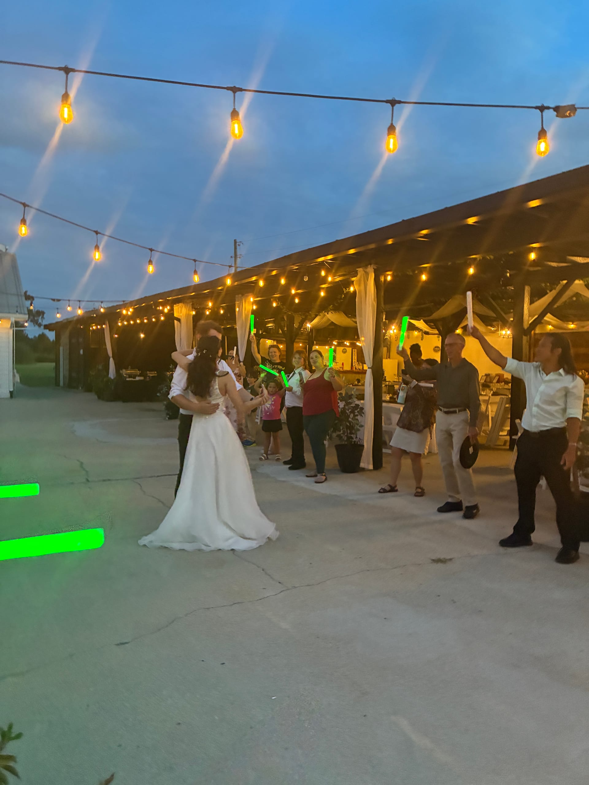 Bride and groom share a romantic first dance outdoors at dusk under warm string lights while wedding guests cheer with glowing green foam sticks at Scarborough House.