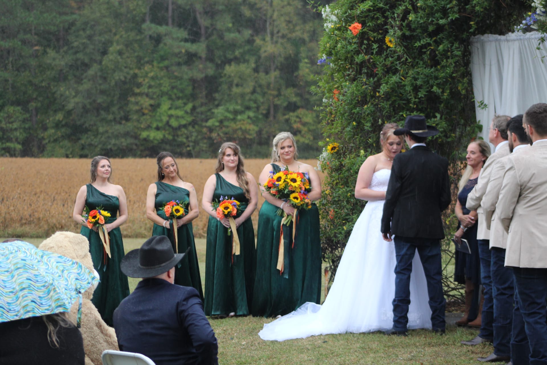 Bride and groom standing at a floral archway on the ceremony lawn at Scarborough House while four bridesmaids in emerald green gowns holding sunflower bouquets look on, with golden farmland fields and lush trees in the background.