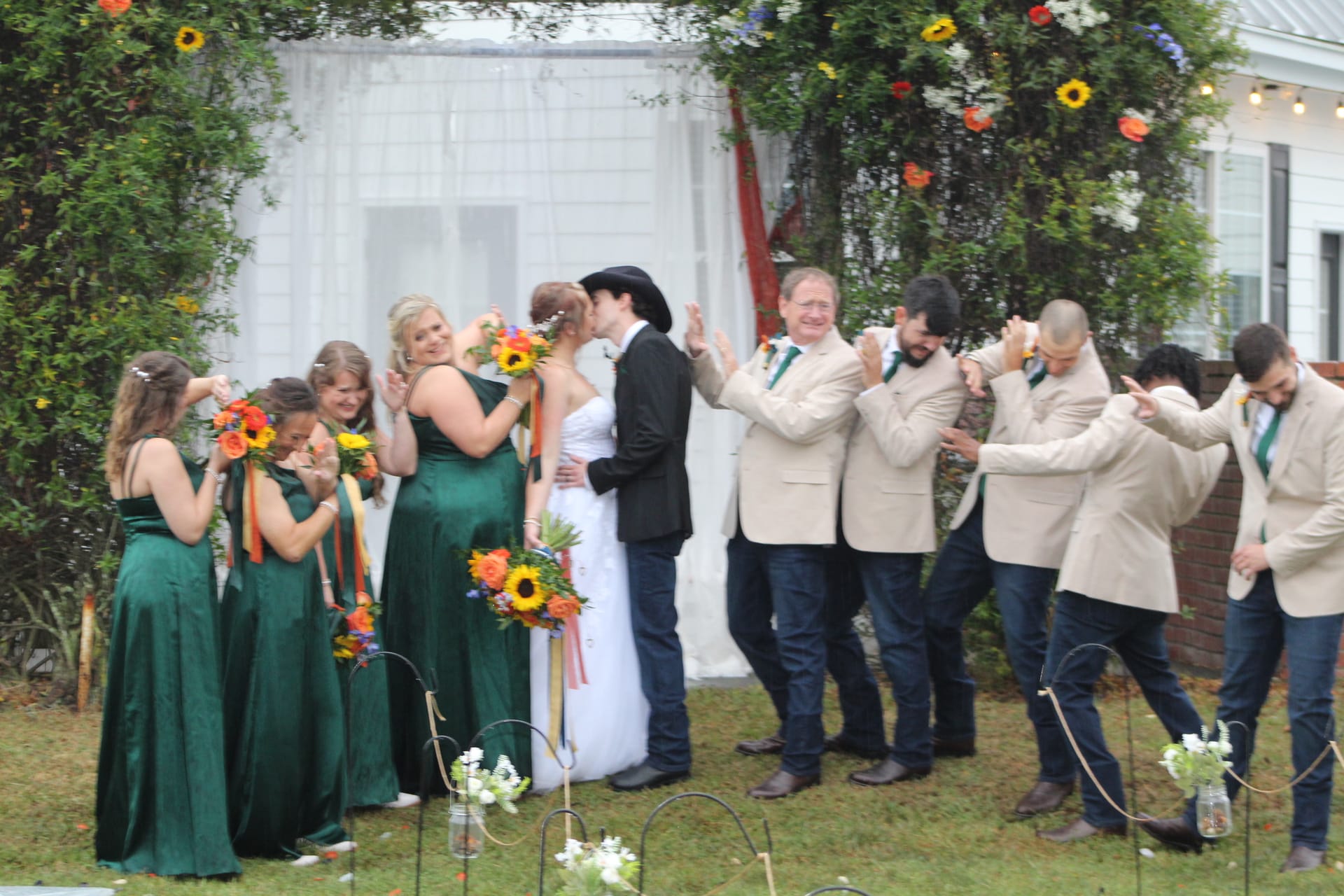 Bride and groom share their first kiss beneath a floral arch at Scarborough House while bridesmaids in emerald gowns and groomsmen in tan blazers celebrate around them on the ceremony lawn.