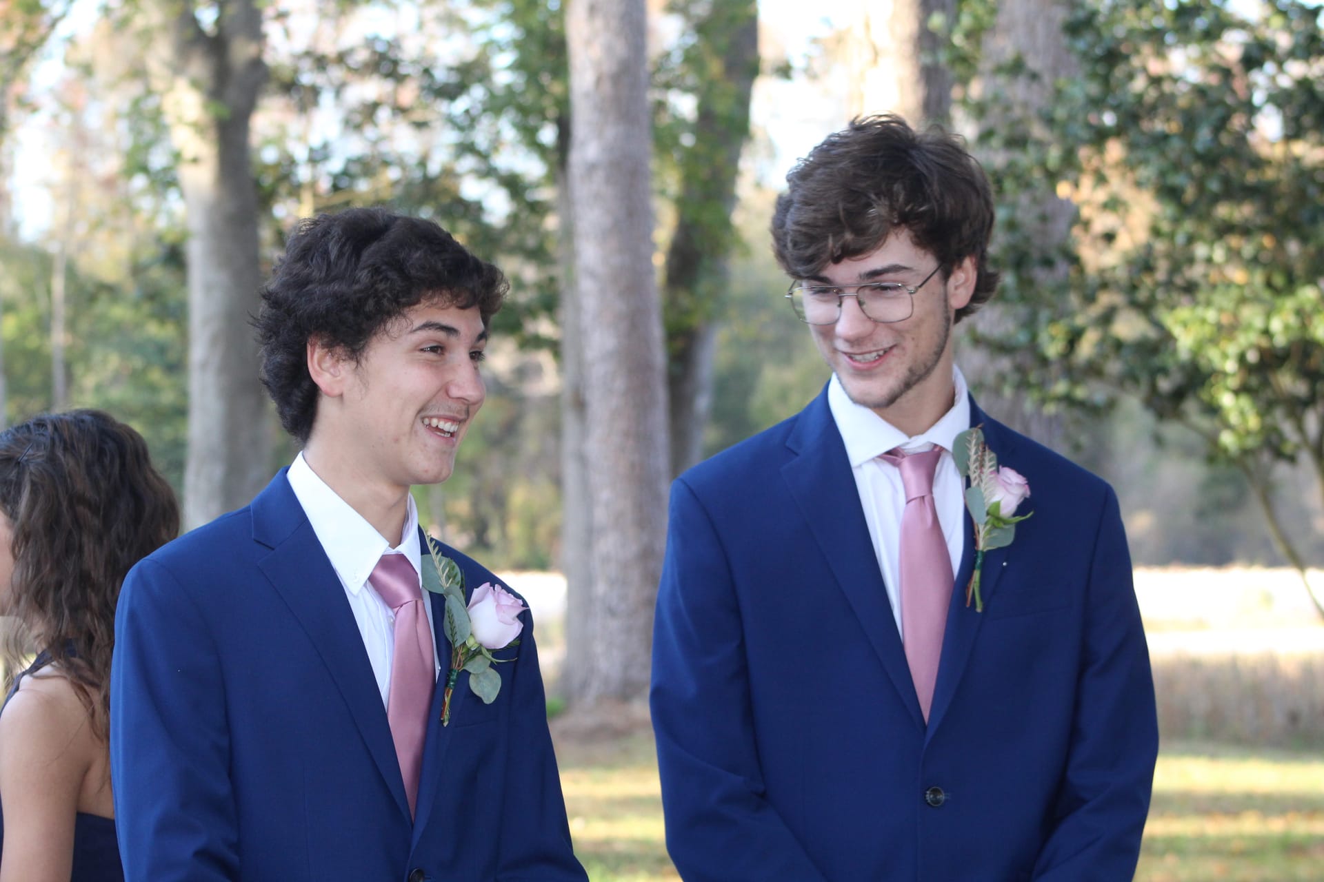 Two young groomsmen in matching navy suits and pink ties smile and laugh together on the ceremony lawn at Scarborough House surrounded by tall oak trees.