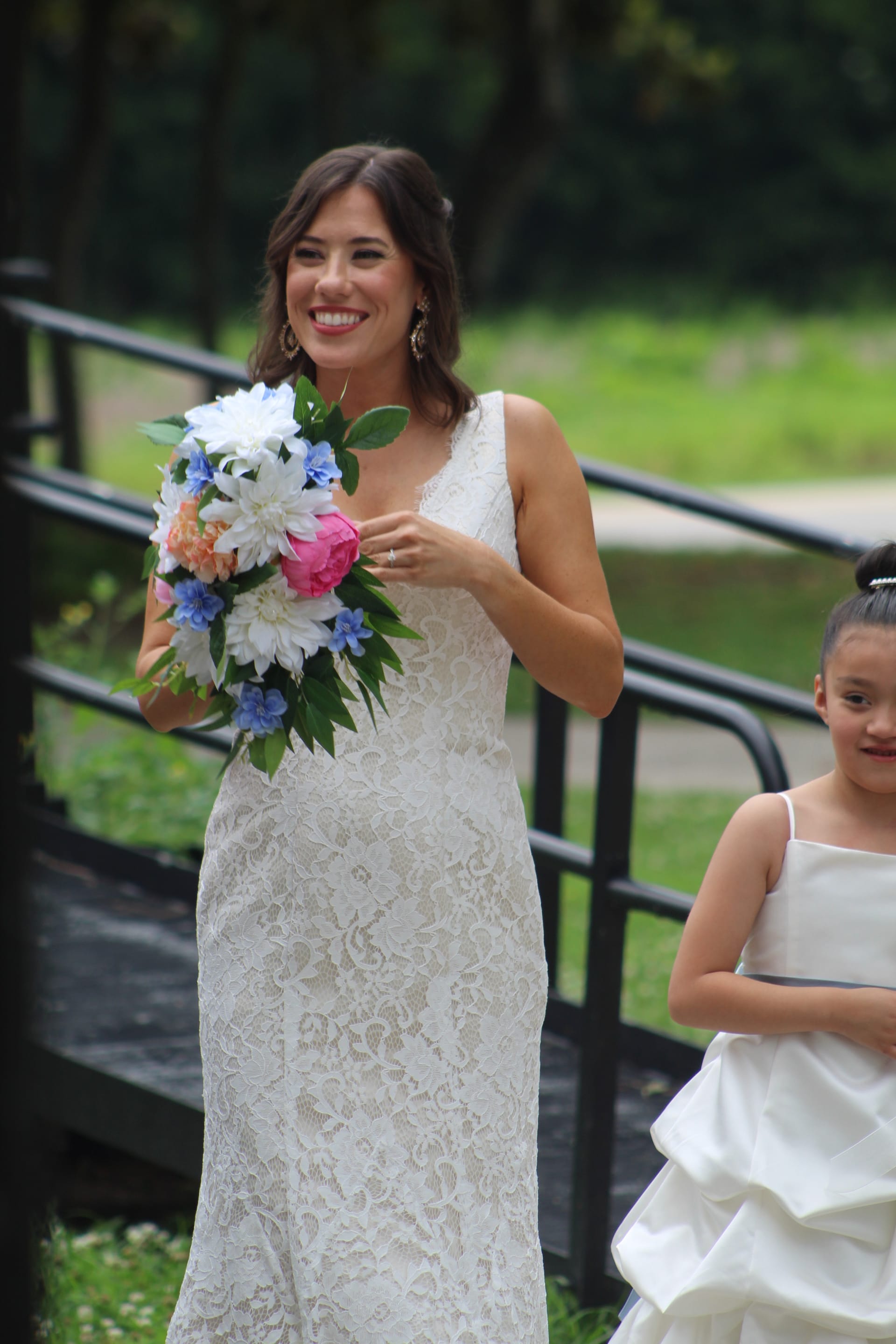Smiling bride in a white lace gown holding a colorful bouquet of pink, white, and blue flowers walks down outdoor steps with a flower girl at a historic estate wedding venue.