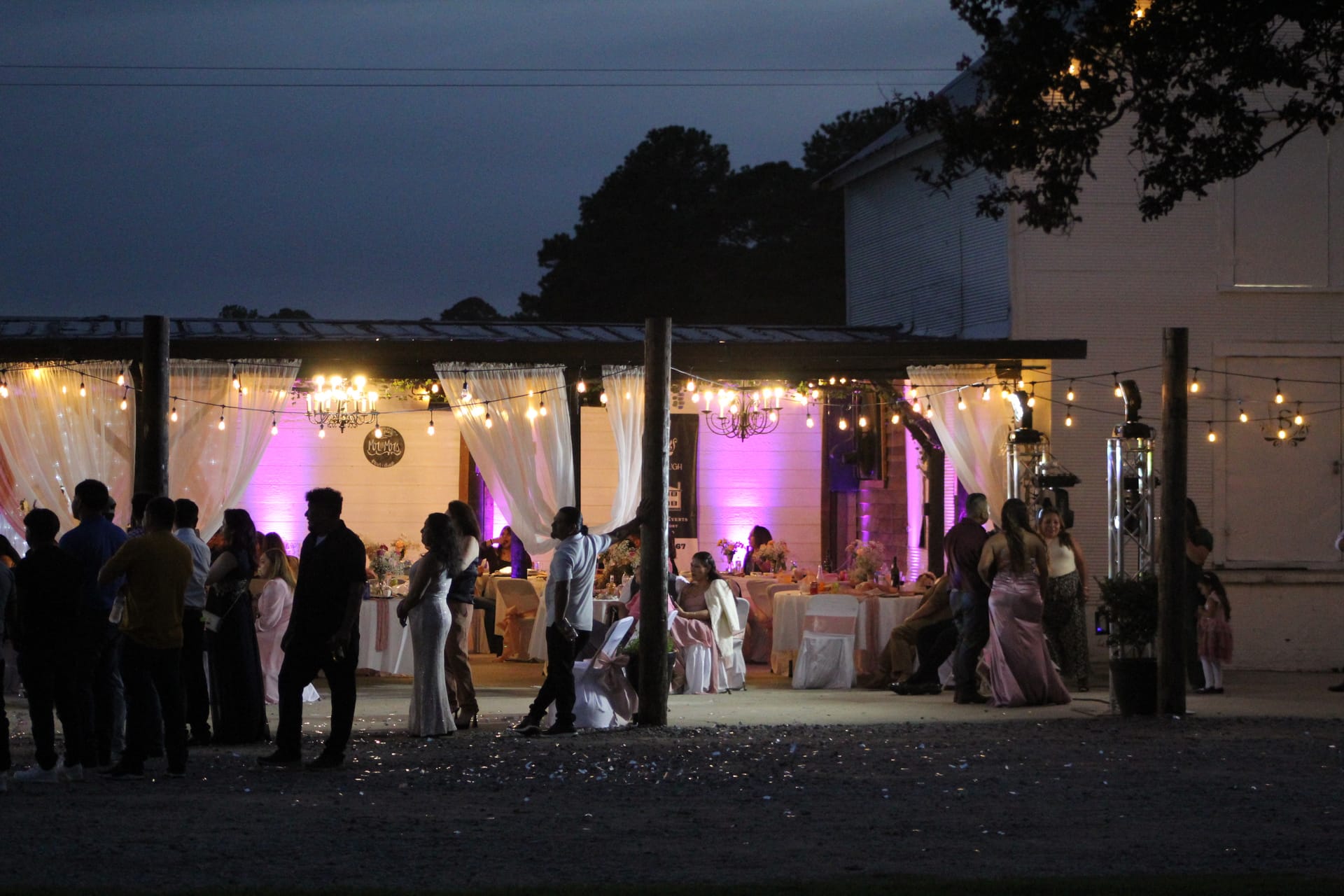 Guests mingle and dance under a warmly lit outdoor pavilion adorned with string lights, chandeliers, white drapery, and purple uplighting at a twilight wedding reception at Scarborough House.