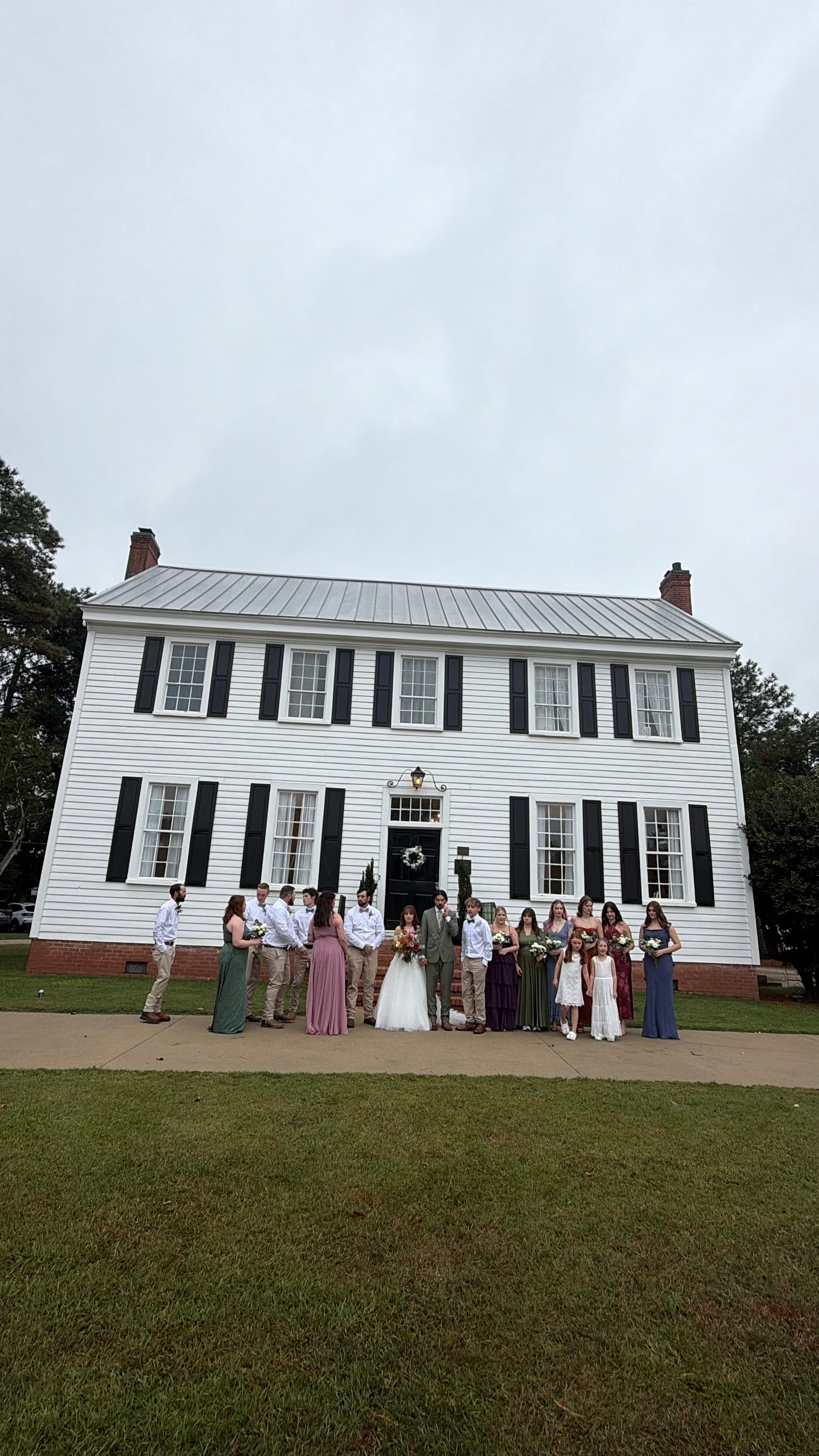 Full wedding party posed in front of the white two-story 1821 Scarborough House manor with black shutters and a wreath on the front door on a lush green lawn.