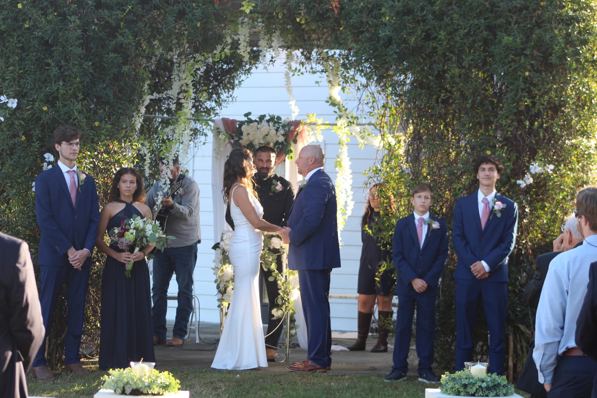 Bride and groom holding hands and exchanging vows on the ceremony lawn at Scarborough House, framed by a lush green arch with white floral accents and attended by their wedding party.