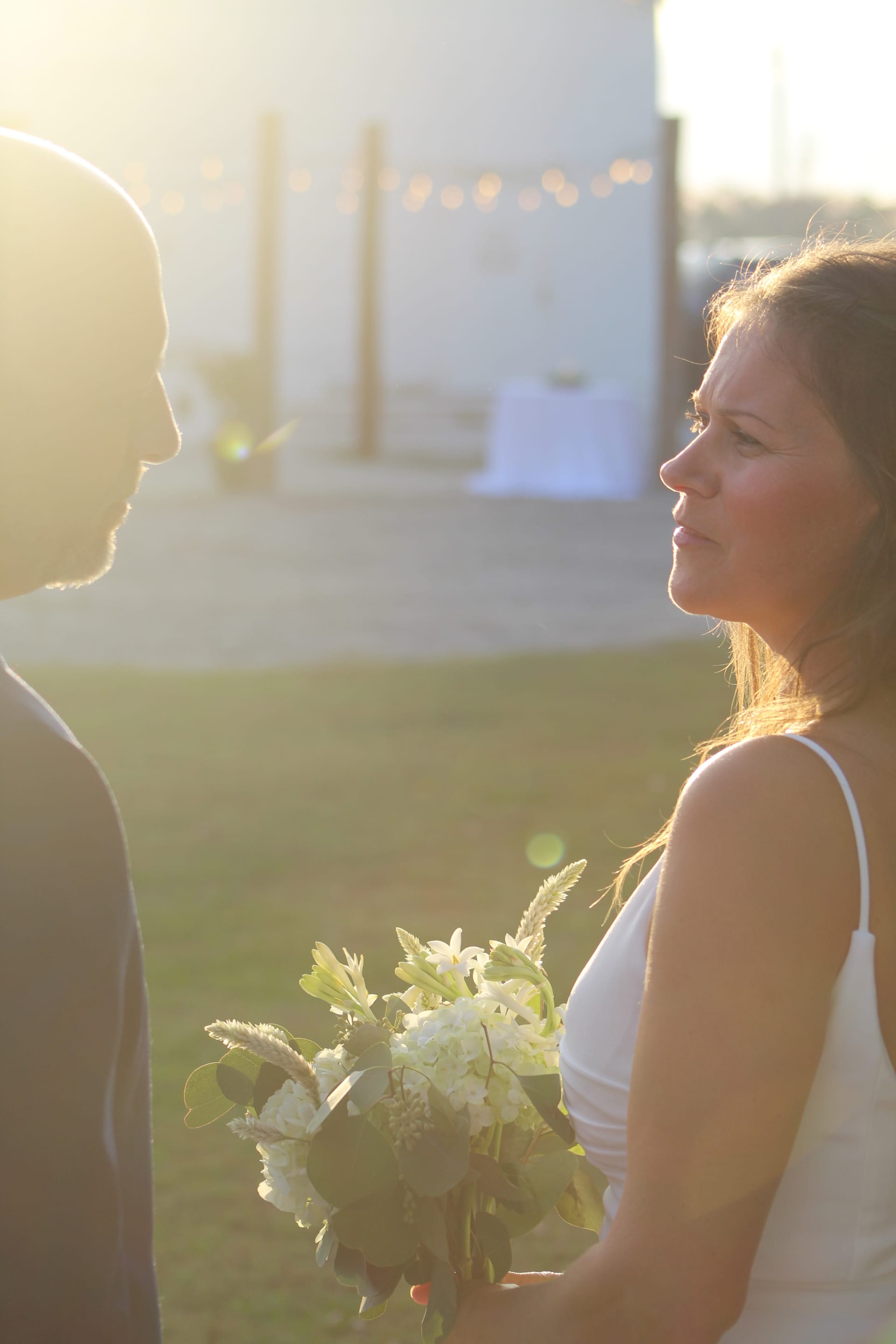 Bride holding a white floral bouquet gazes at her groom in warm golden sunset light on the ceremony lawn at Scarborough House estate.