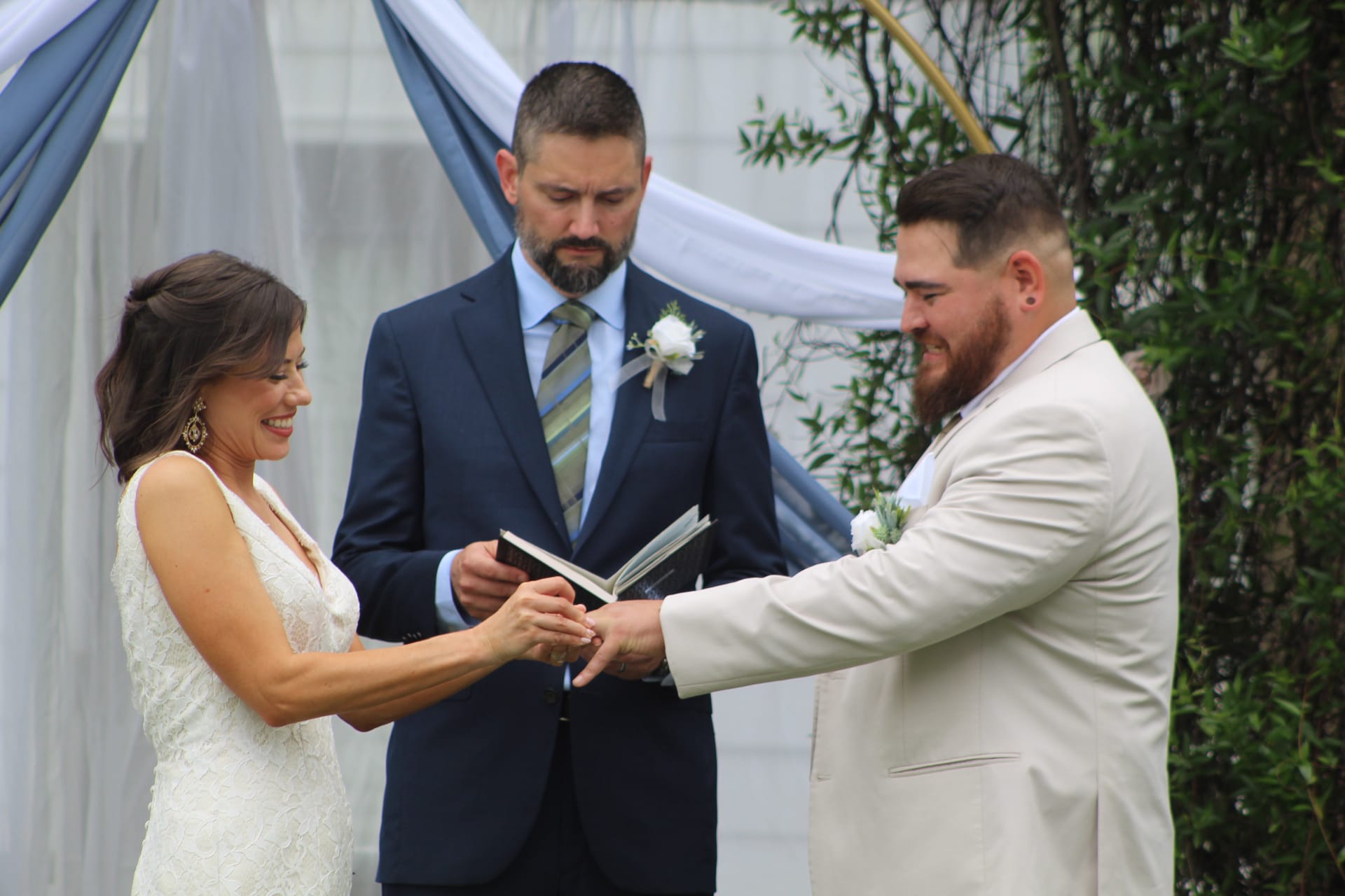 Bride and groom exchange rings during an outdoor wedding ceremony at Scarborough House with a draped arch and officiant behind them.