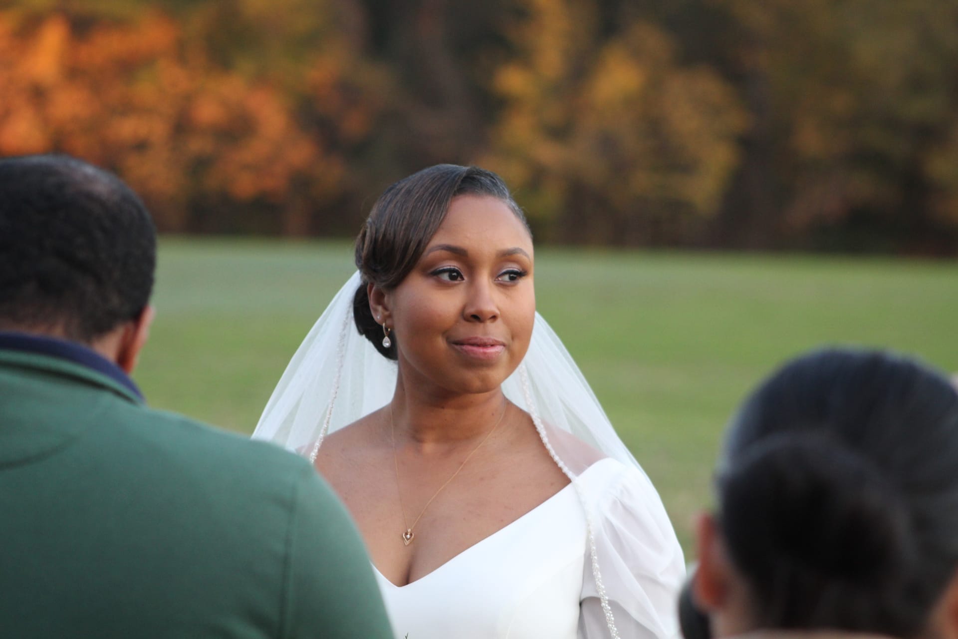 Bride in a white gown and veil gazes forward with a tender expression during an outdoor autumn wedding ceremony at Scarborough House, with golden foliage and green fields in the background.