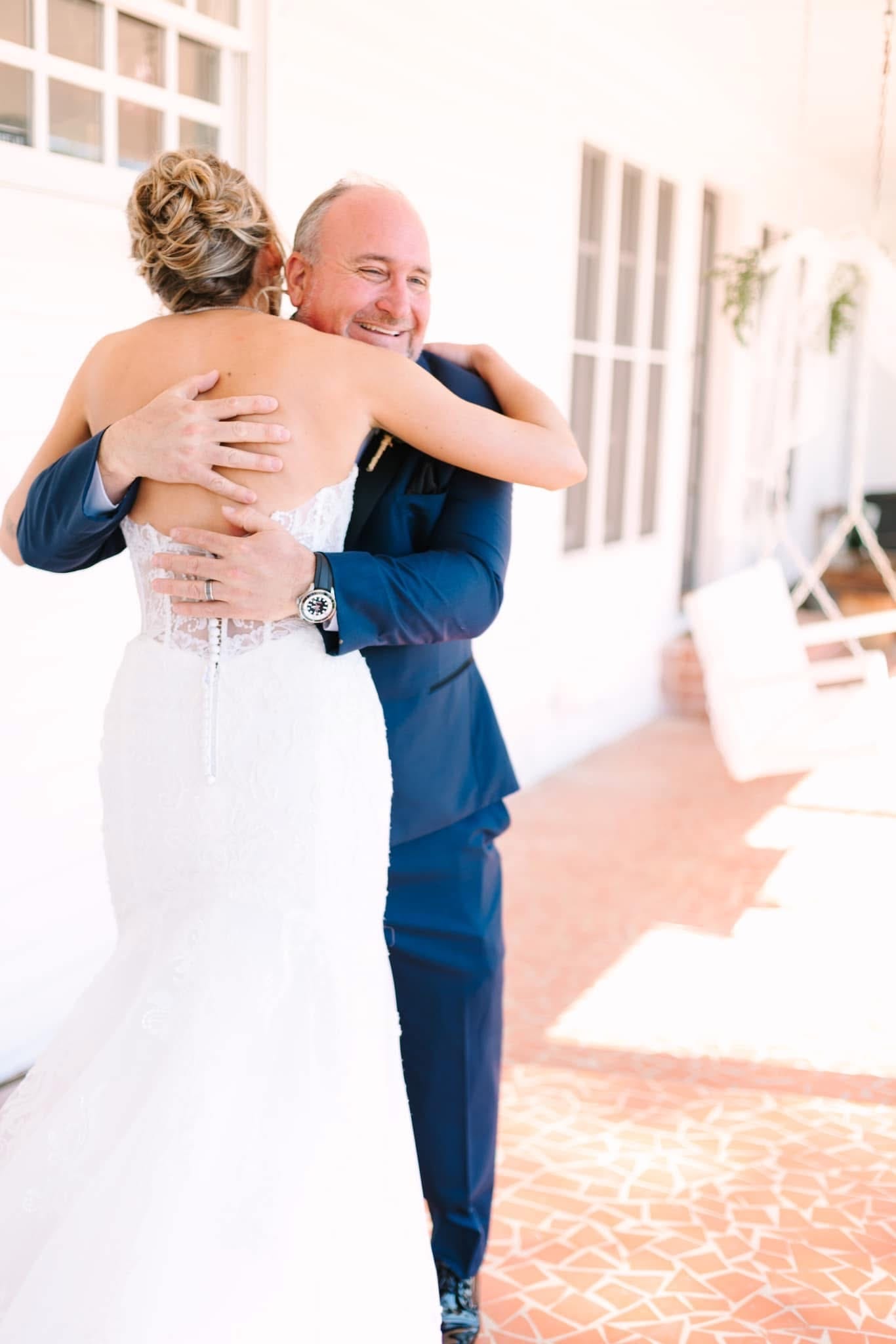 A bride in a fitted lace gown embraces a smiling man in a navy suit on the sunlit porch of the historic Scarborough House estate.