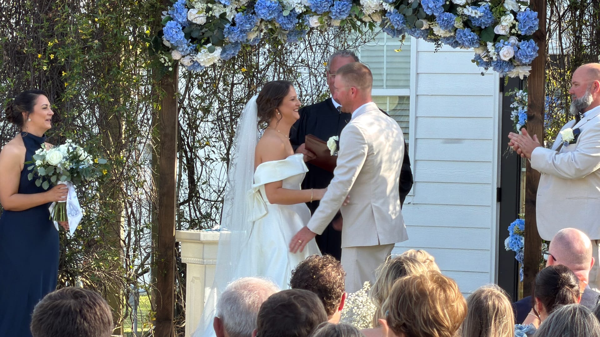 Bride and groom share a joyful moment during their outdoor wedding ceremony beneath a blue and white hydrangea floral arch at Scarborough House historic estate.