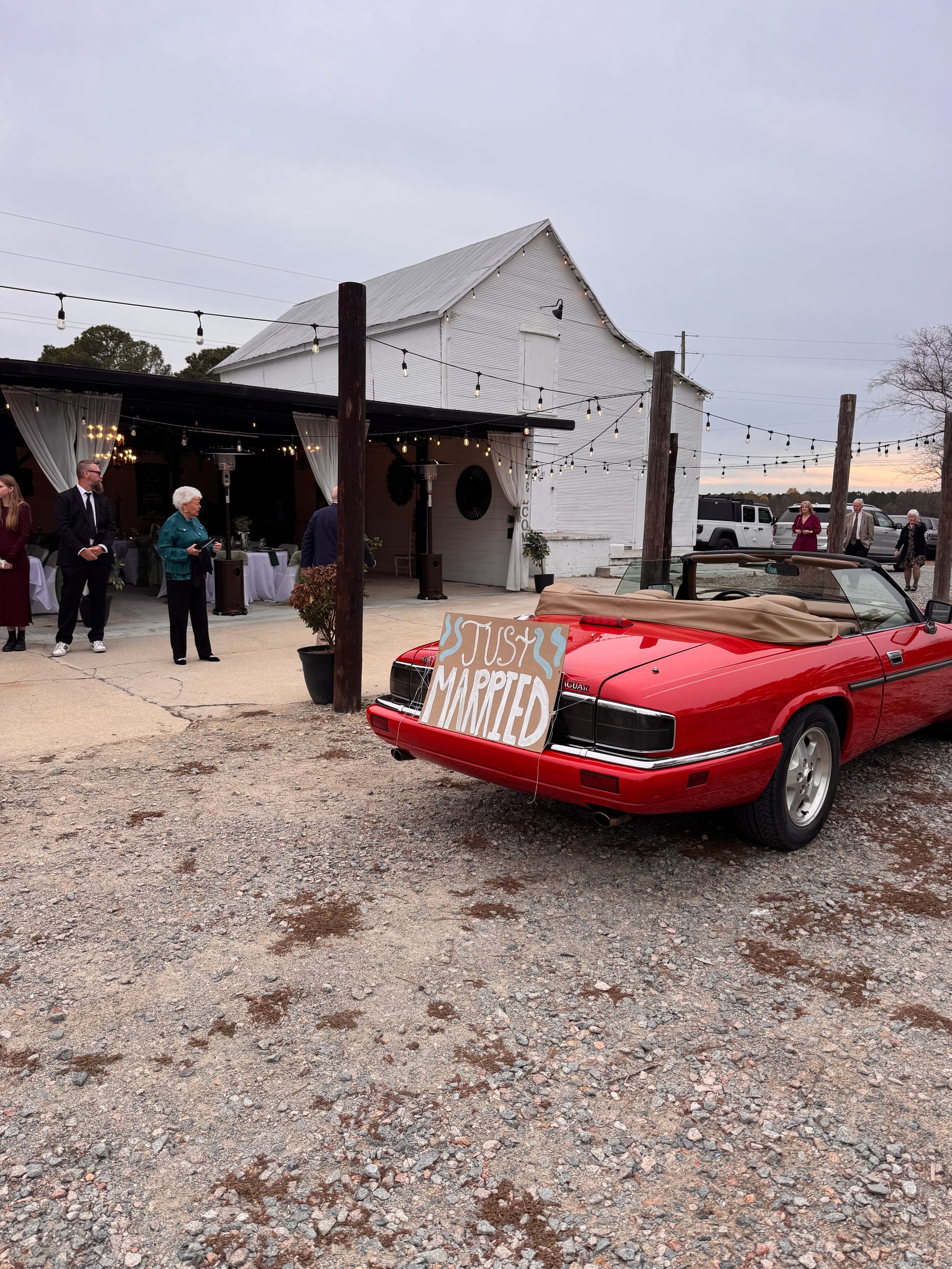 A red convertible decorated with a Just Married sign parked outside a white barn wedding venue strung with bistro lights at dusk.