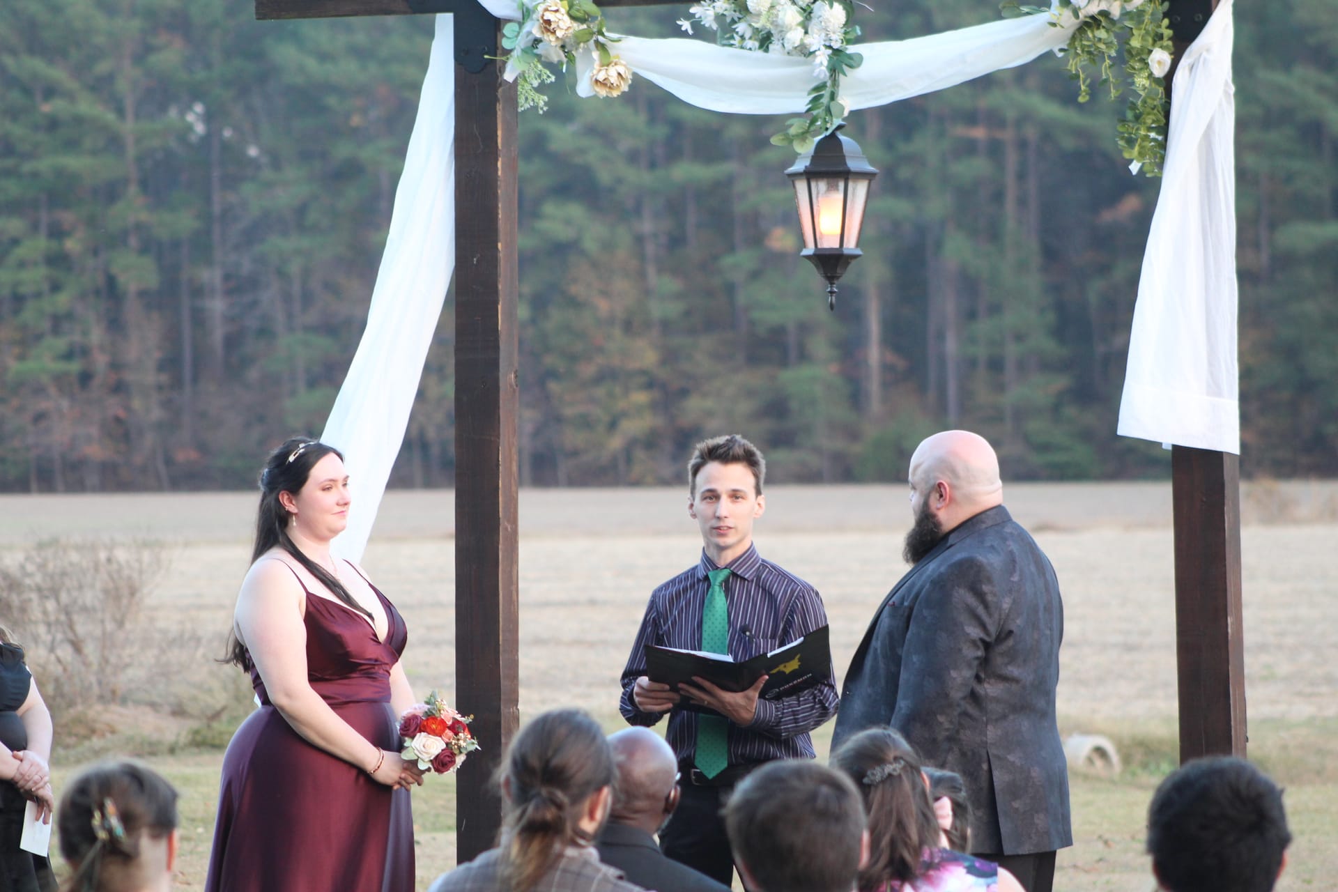 Bride in a burgundy gown and groom in a dark suit exchange vows with an officiant under a white-draped wooden arch adorned with floral arrangements and a hanging lantern on the ceremony lawn at Scarborough House.