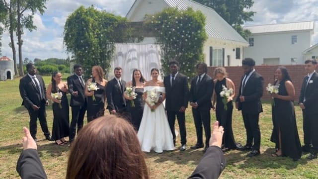 Bride and groom stand centered beneath a draped floral arch surrounded by their wedding party dressed in black on the ceremony lawn at Scarborough House historic estate.
