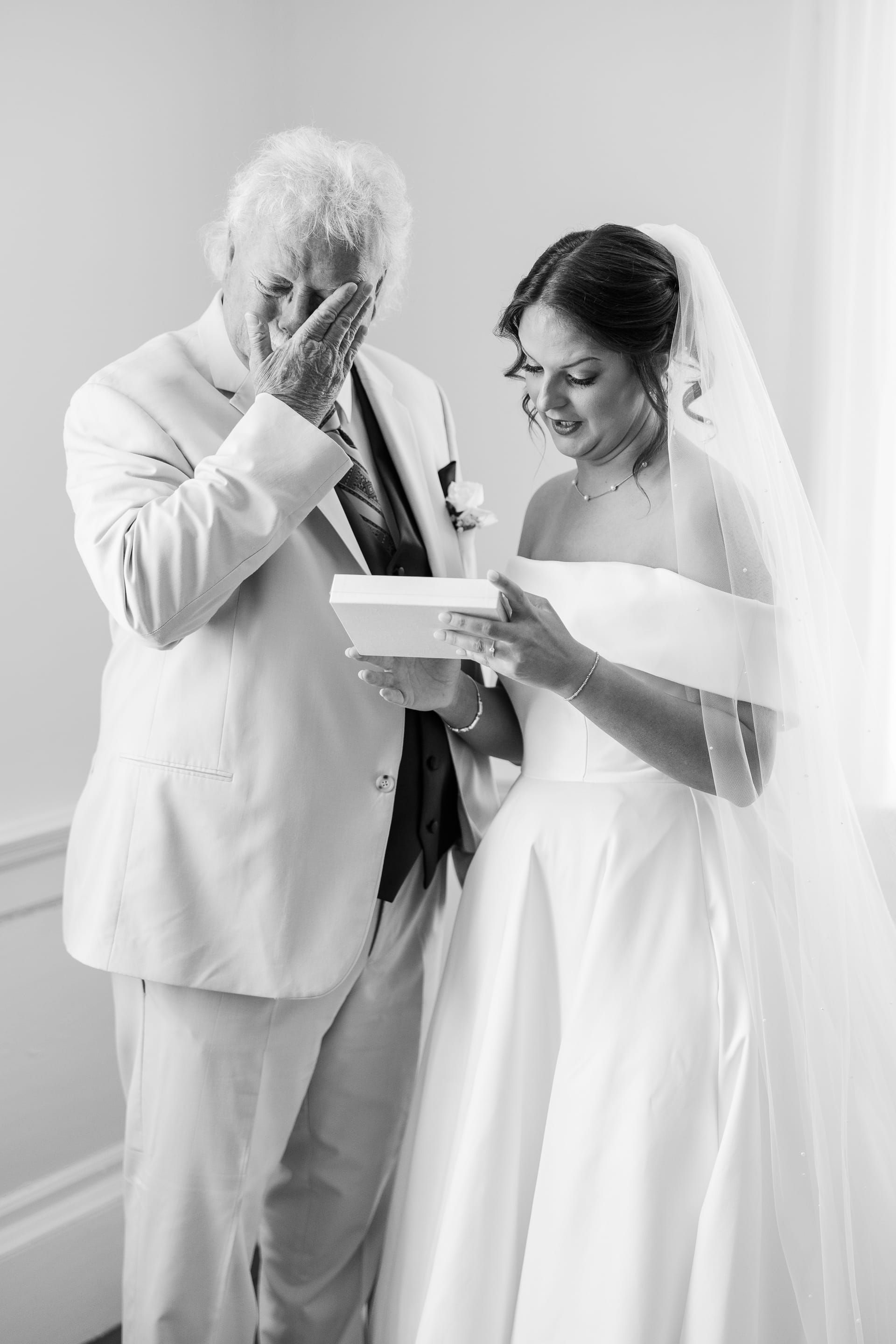 A bride in a white gown and veil shares an emotional moment with an older gentleman in a suit as they look at a gift box together, both visibly moved to tears.