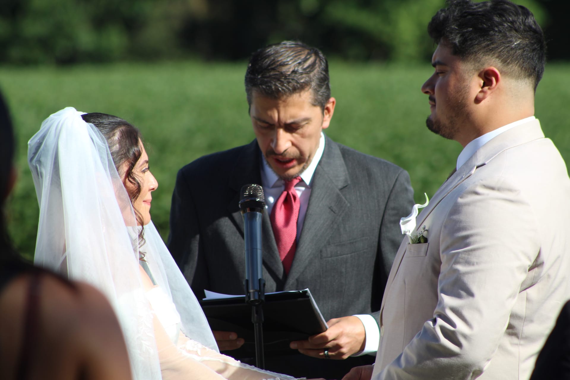 Bride in white veil and groom in tan suit exchange vows before an officiant in a gray suit at an outdoor wedding ceremony surrounded by greenery.