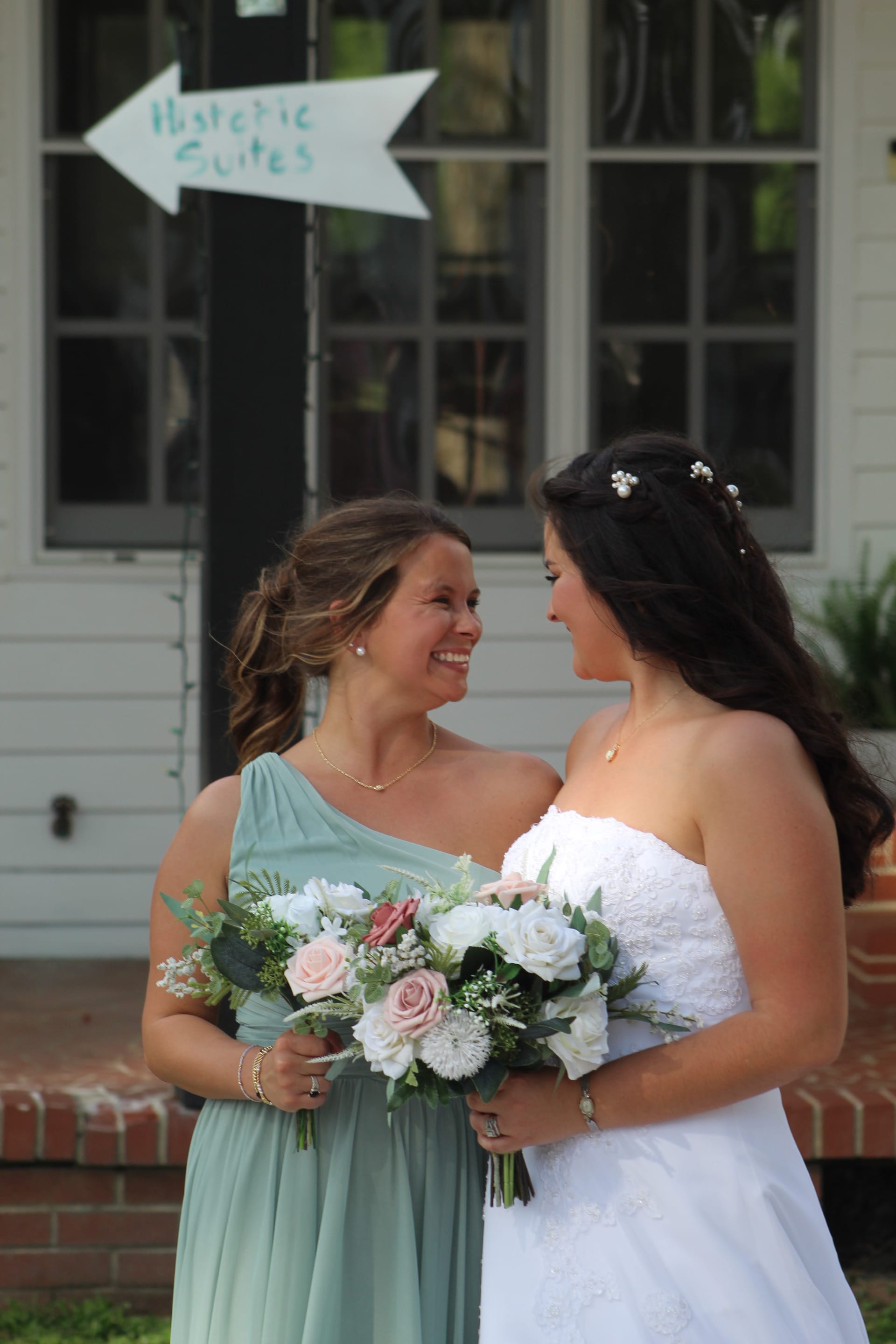 Bride in a strapless white gown and bridesmaid in a sage one-shoulder dress share a joyful smile while holding blush and white floral bouquets in front of the historic Scarborough House manor.