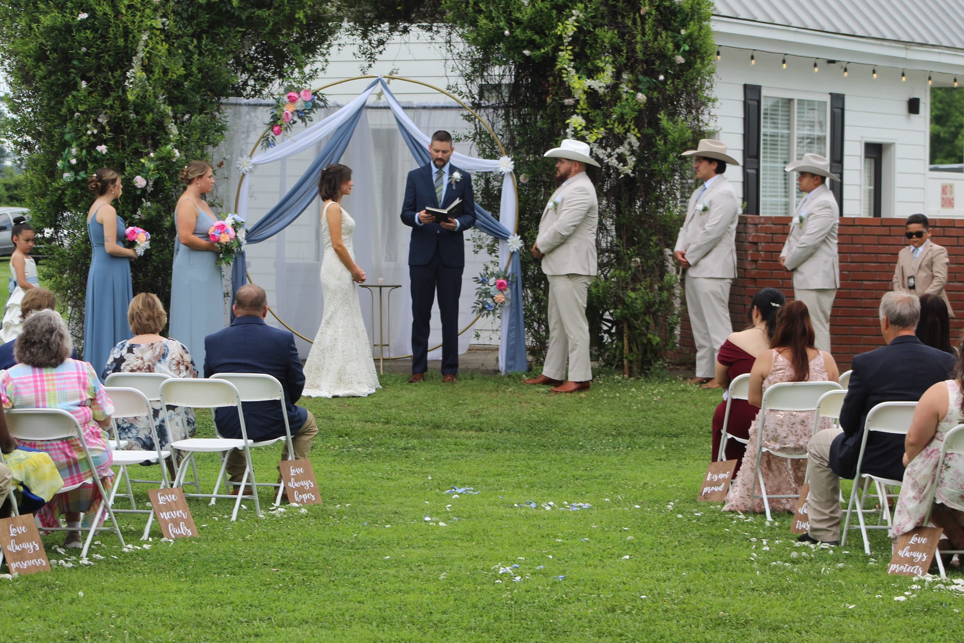 Bride and groom exchange vows beneath a blue draped circular arch adorned with florals on the ceremony lawn at Scarborough House, surrounded by bridesmaids in dusty blue, groomsmen in tan suits, and seated guests.