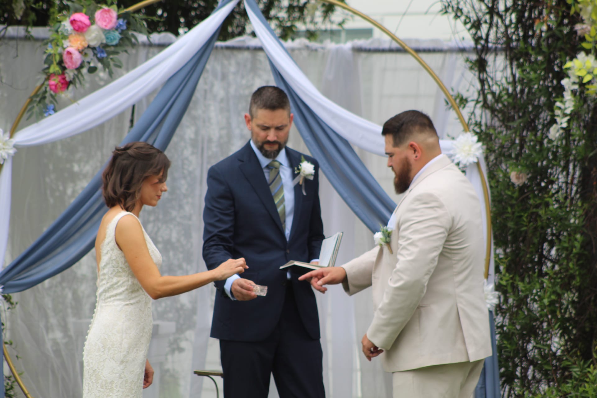 Bride and groom exchange rings before an officiant beneath a gold circular arch adorned with colorful florals and dusty blue draped fabric during an outdoor wedding ceremony.