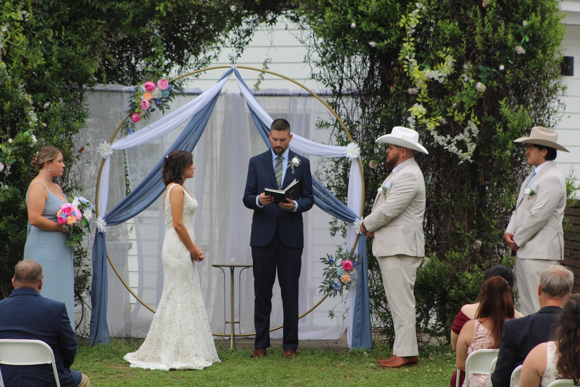 Bride and groom exchange vows before a gold circular arch draped in blue fabric and fresh flowers on the ceremony lawn at Scarborough House historic estate.