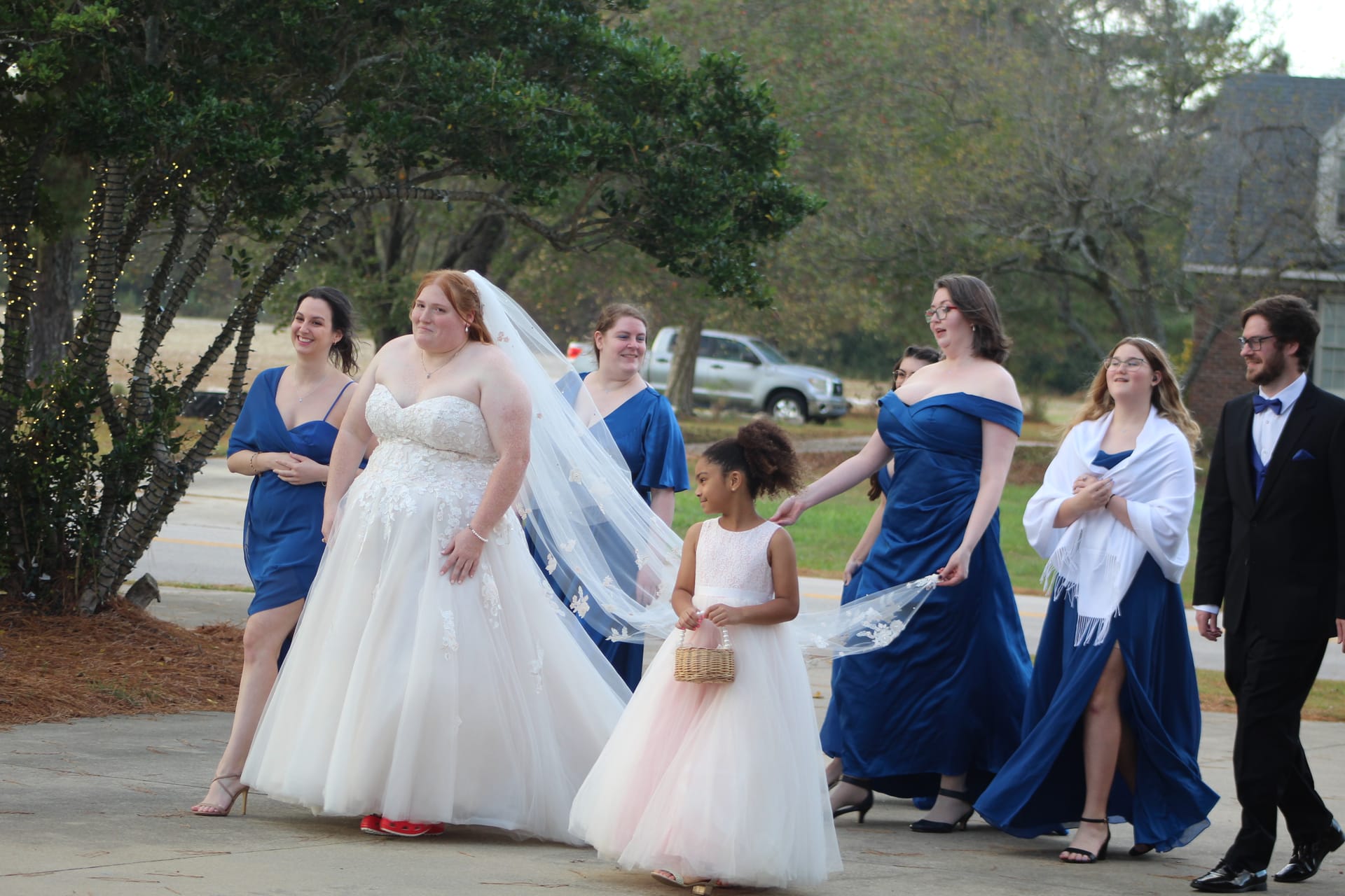 Bride in a lace ballgown with veil carried by a young flower girl walks alongside bridesmaids in royal blue dresses on the grounds of a historic estate wedding venue.