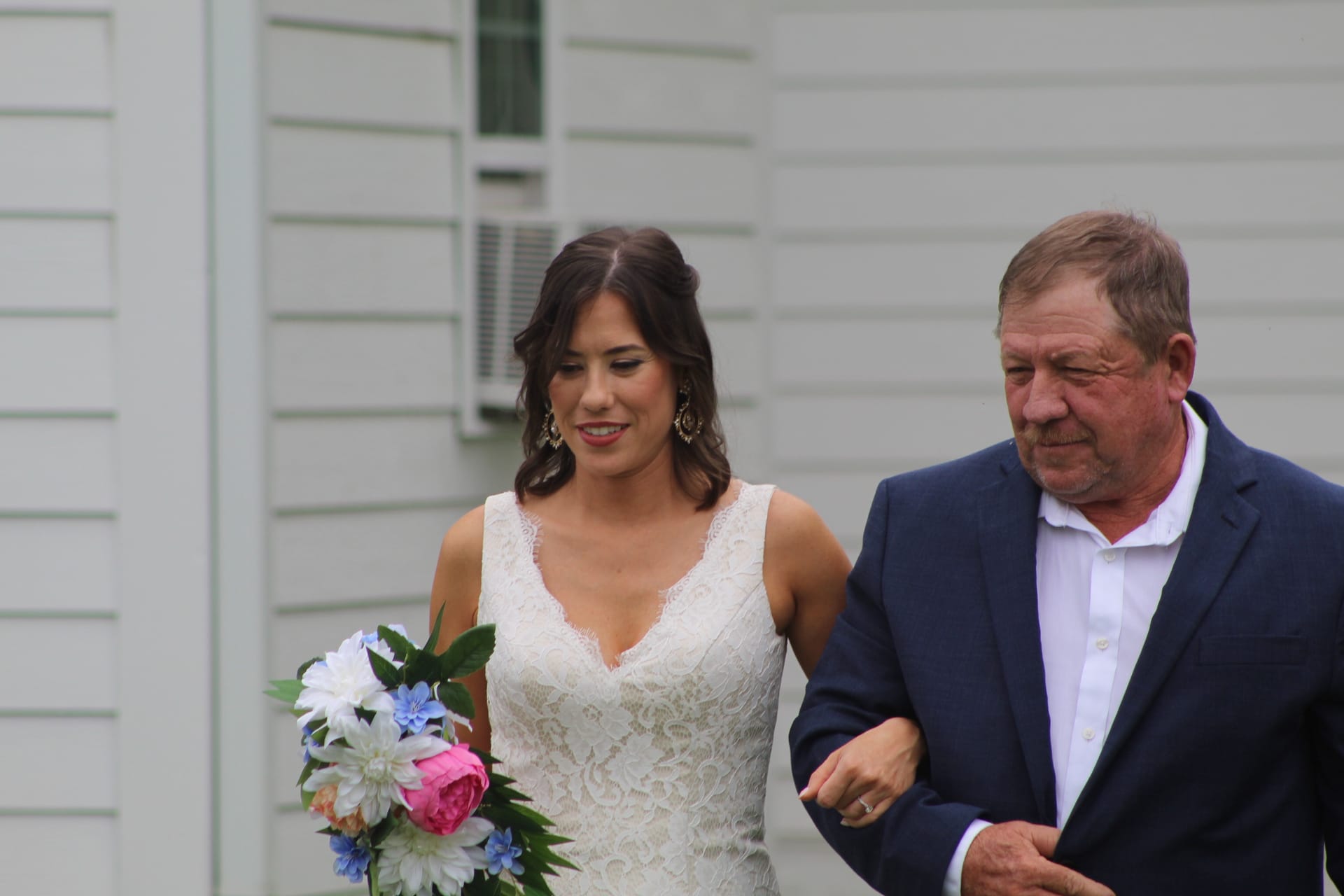 Bride in a lace wedding gown holding a pink and white bouquet walks arm in arm with an older gentleman in a navy blazer past the exterior of the historic Scarborough House estate.