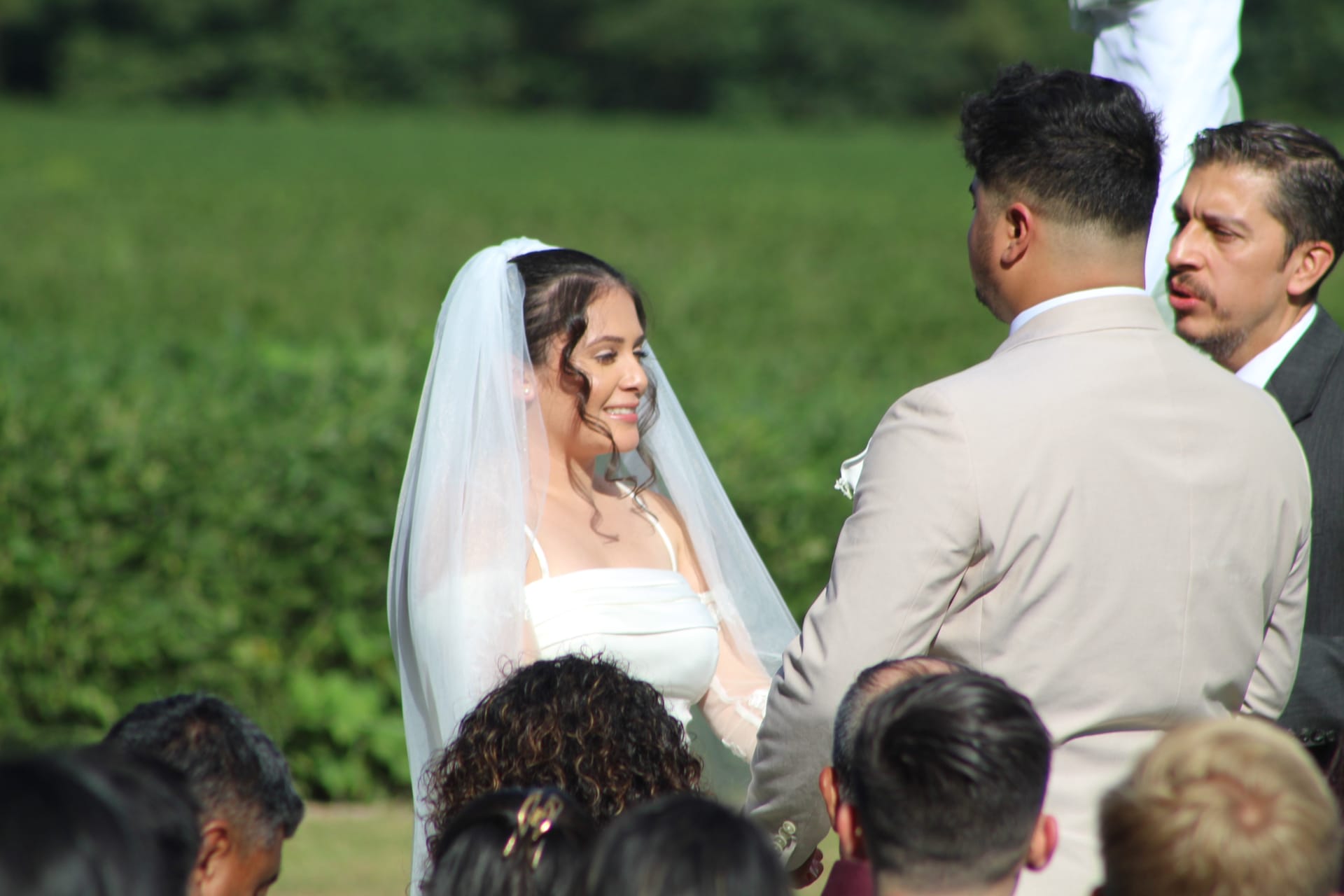 Smiling bride in white gown and veil exchanges vows with groom in tan suit on the ceremony lawn at Scarborough House estate surrounded by lush green fields.