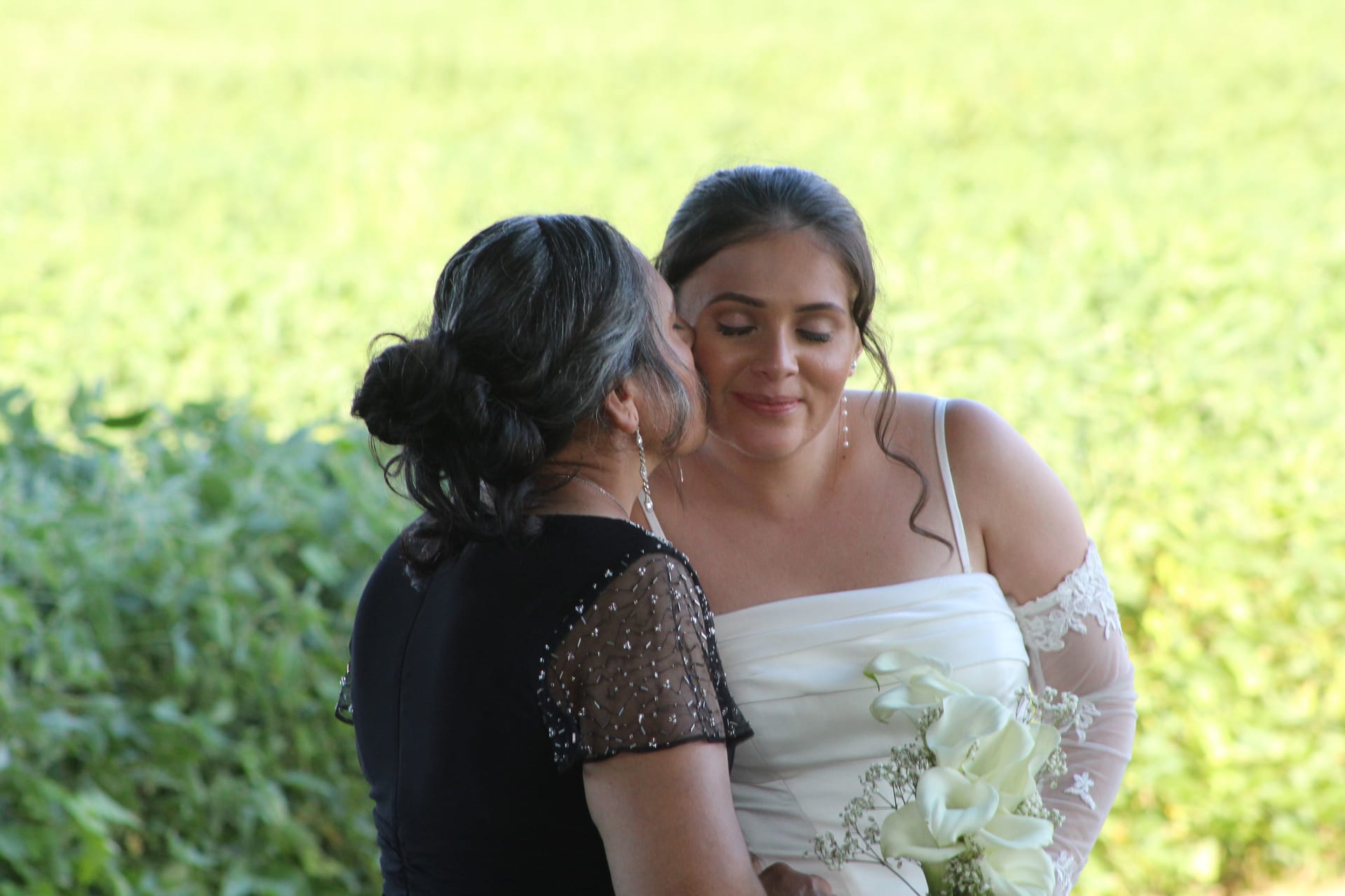A woman in a black dress tenderly kisses a smiling bride on the cheek as she holds a white calla lily bouquet against a lush green garden backdrop at Scarborough House.