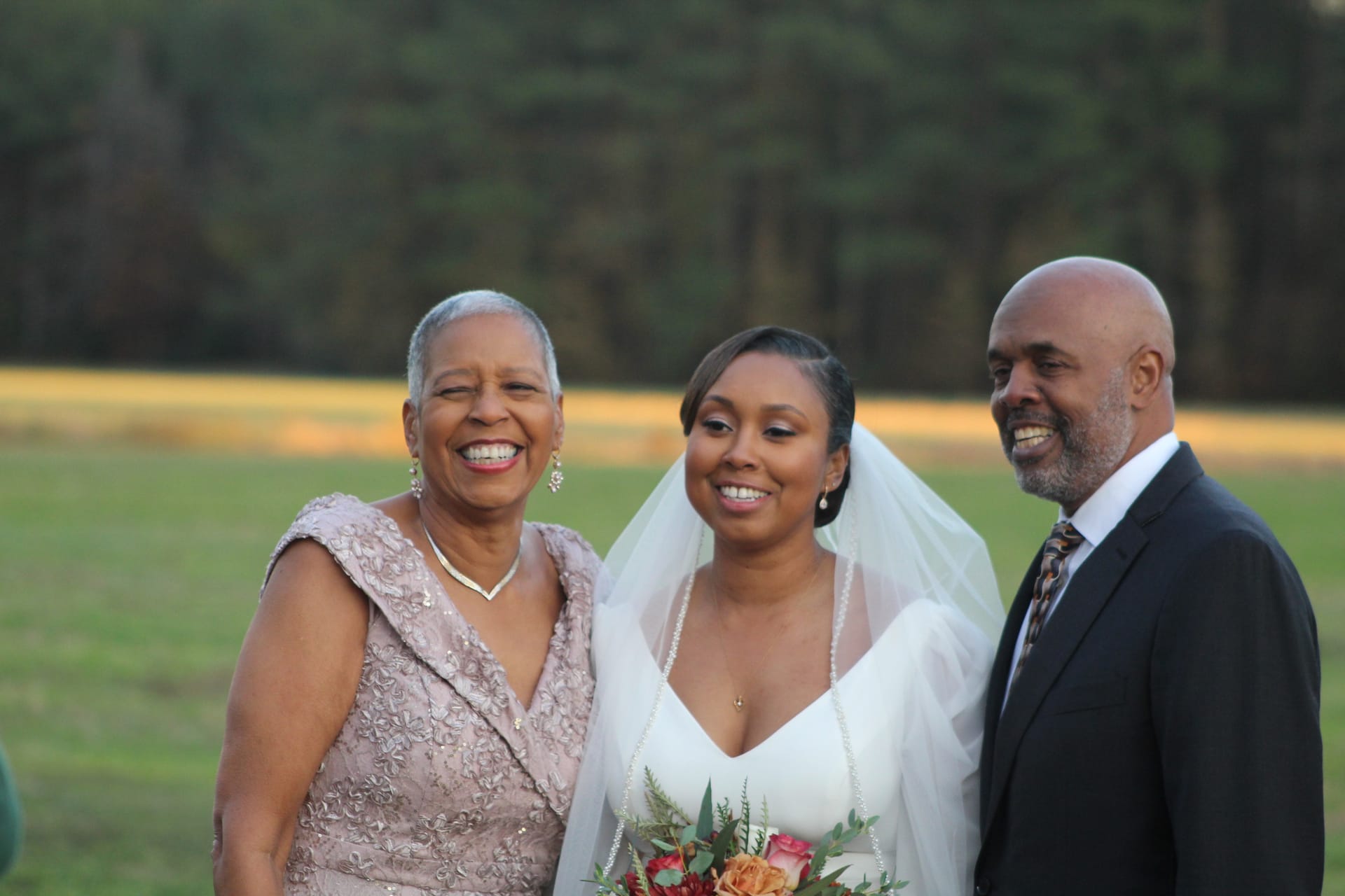 A radiant bride in a white gown and veil holds a floral bouquet while smiling with her elegantly dressed mother and suited father on the sun-kissed open fields of Scarborough House.