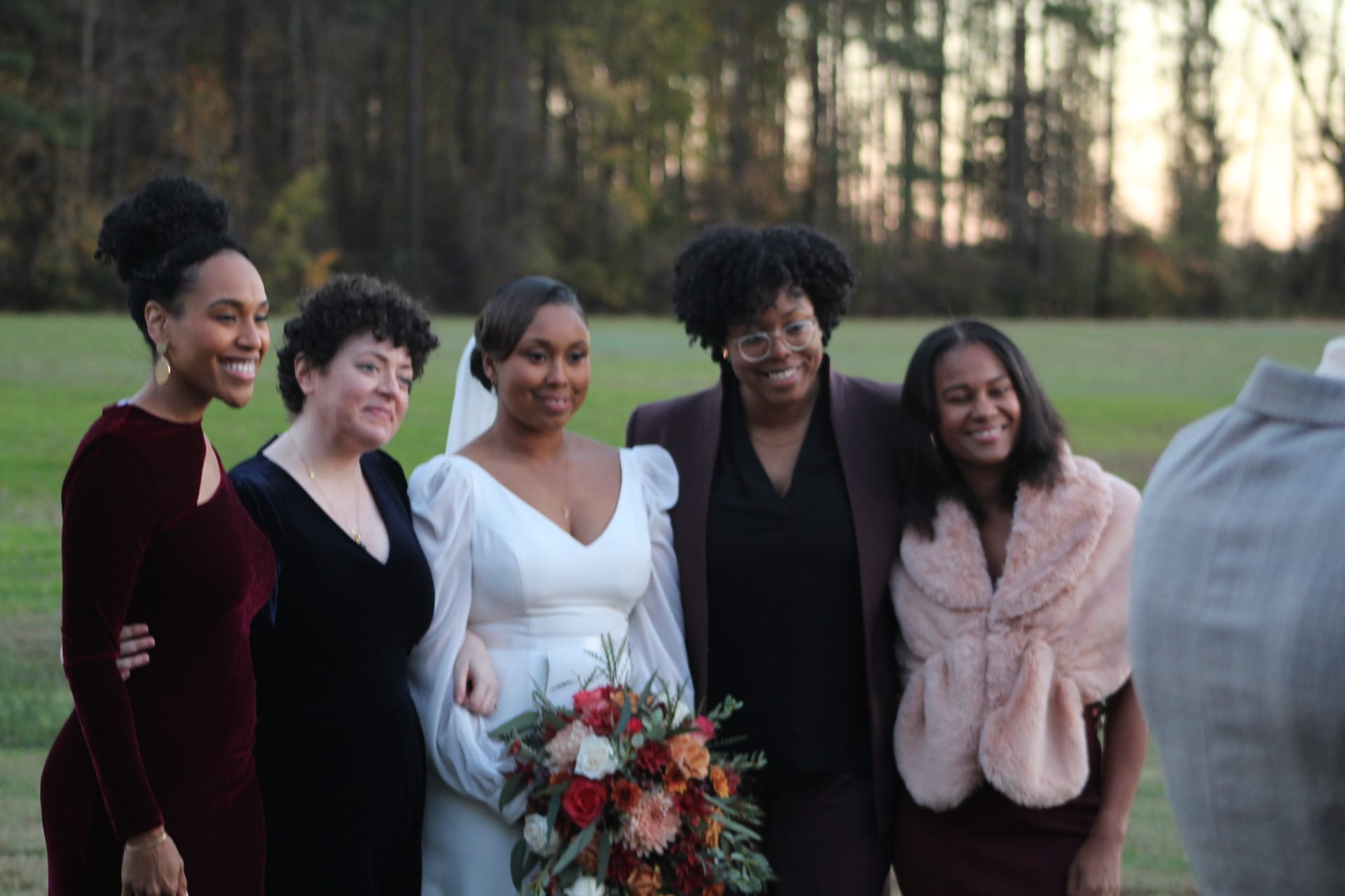 Bride in a white long-sleeve gown holding a colorful fall bouquet poses and smiles with four guests on the ceremony lawn surrounded by trees at Scarborough House.
