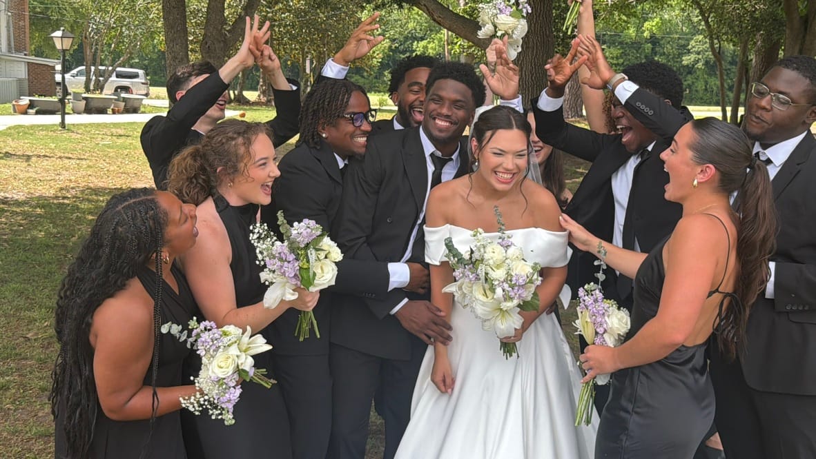 Bride in off-shoulder white gown and groom in black suit laugh joyfully surrounded by an exuberant wedding party holding white and lavender bouquets beneath oak trees at Scarborough House.
