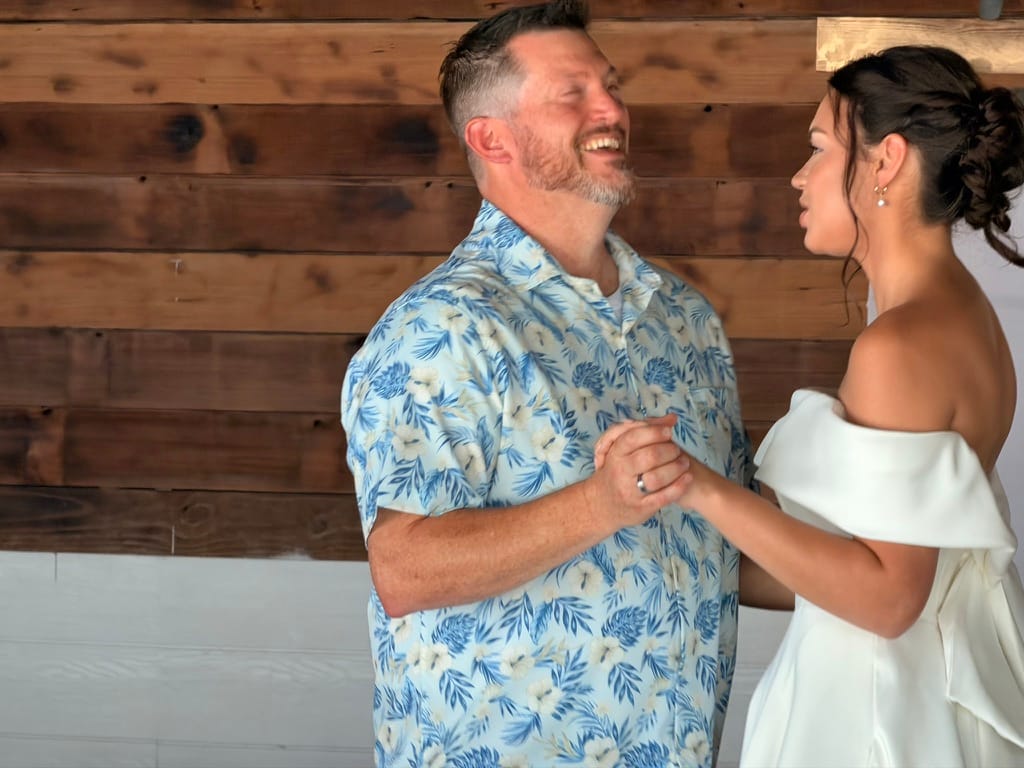 Groom in a blue floral shirt laughs joyfully while holding hands with his bride in an off-shoulder white gown during their first dance at Scarborough House.