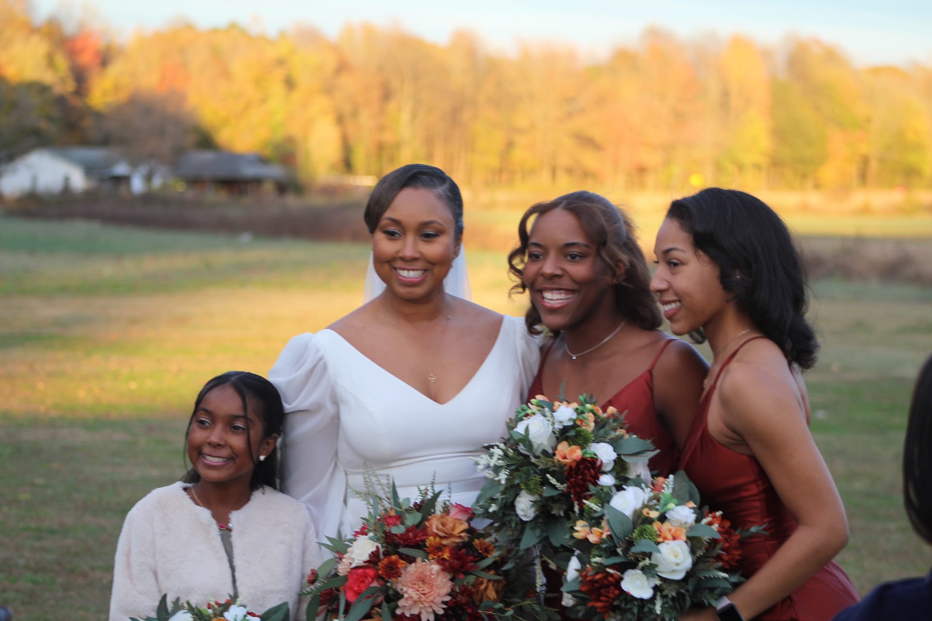 Bride in white gown and veil poses with bridesmaids in burgundy dresses and a young flower girl holding autumn bouquets against a backdrop of golden fall foliage and open farmland fields at Scarborough House.