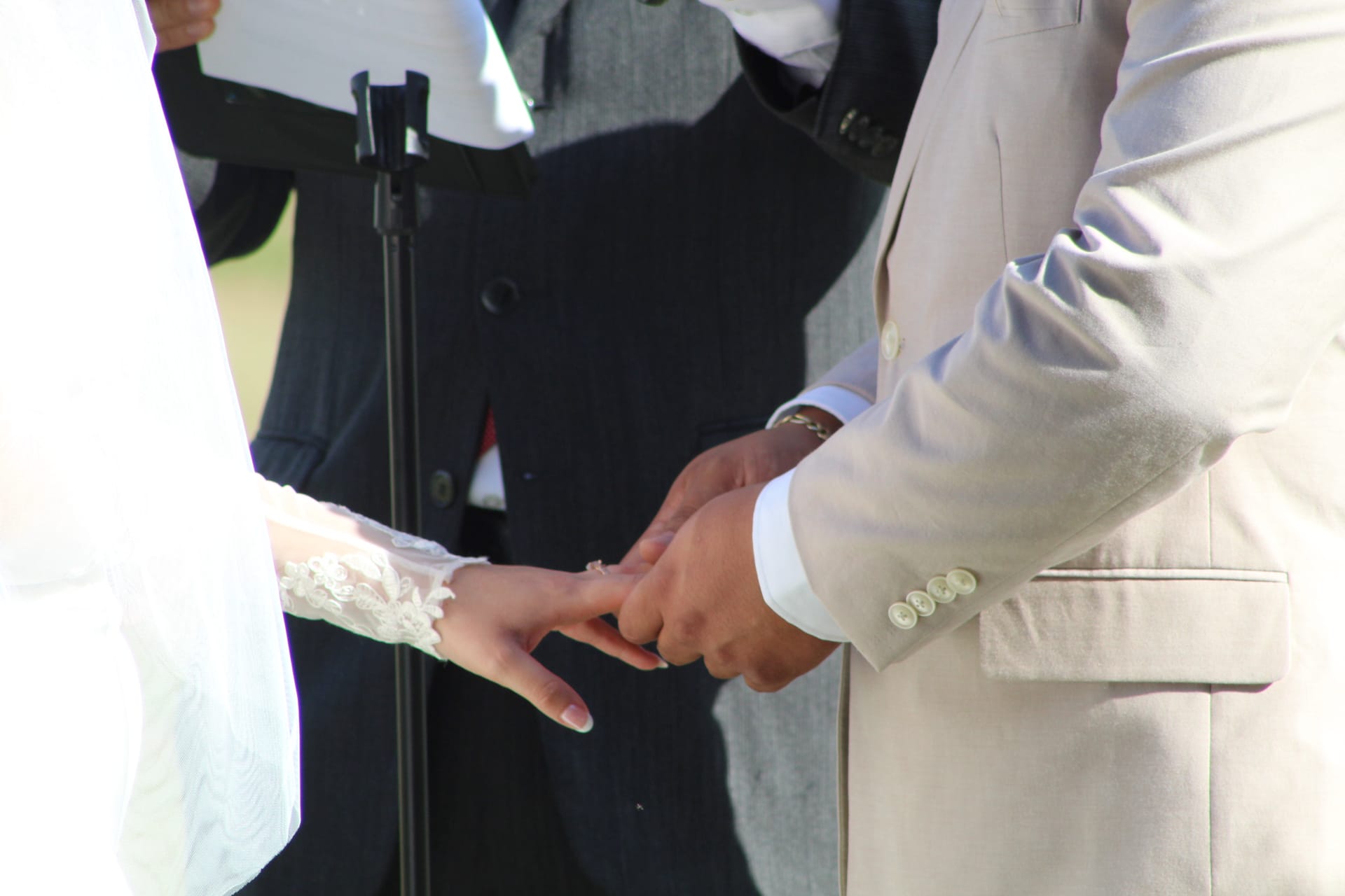 Groom in an ivory suit places a wedding ring on his brides lace-sleeved hand during an outdoor ceremony at Scarborough House.