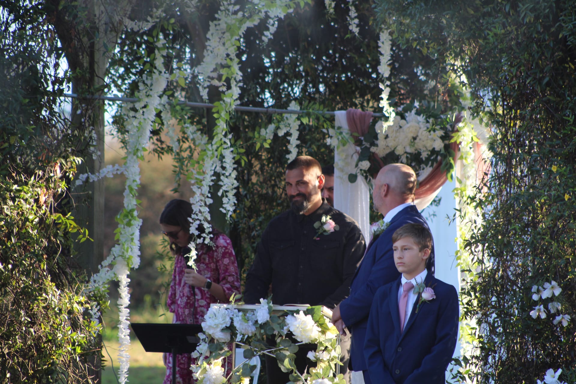Groom smiling joyfully beneath a lush floral arch adorned with hanging white flowers during an outdoor garden wedding ceremony at Scarborough House.
