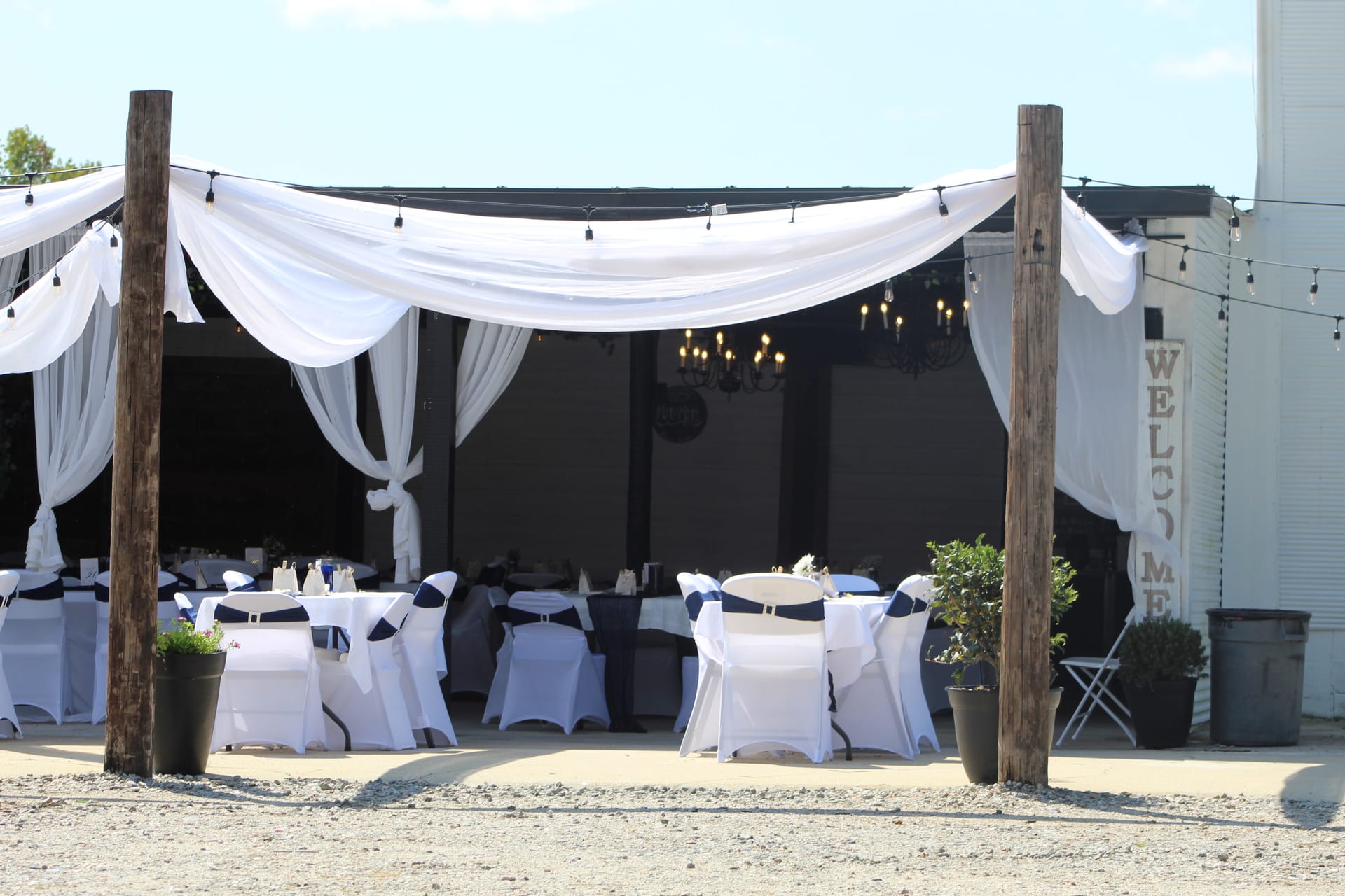 Elegant outdoor wedding reception setup at Scarborough House featuring white draped fabric over rustic wooden posts, navy-sashed white chair covers, string lights, and a Welcome sign in the background.