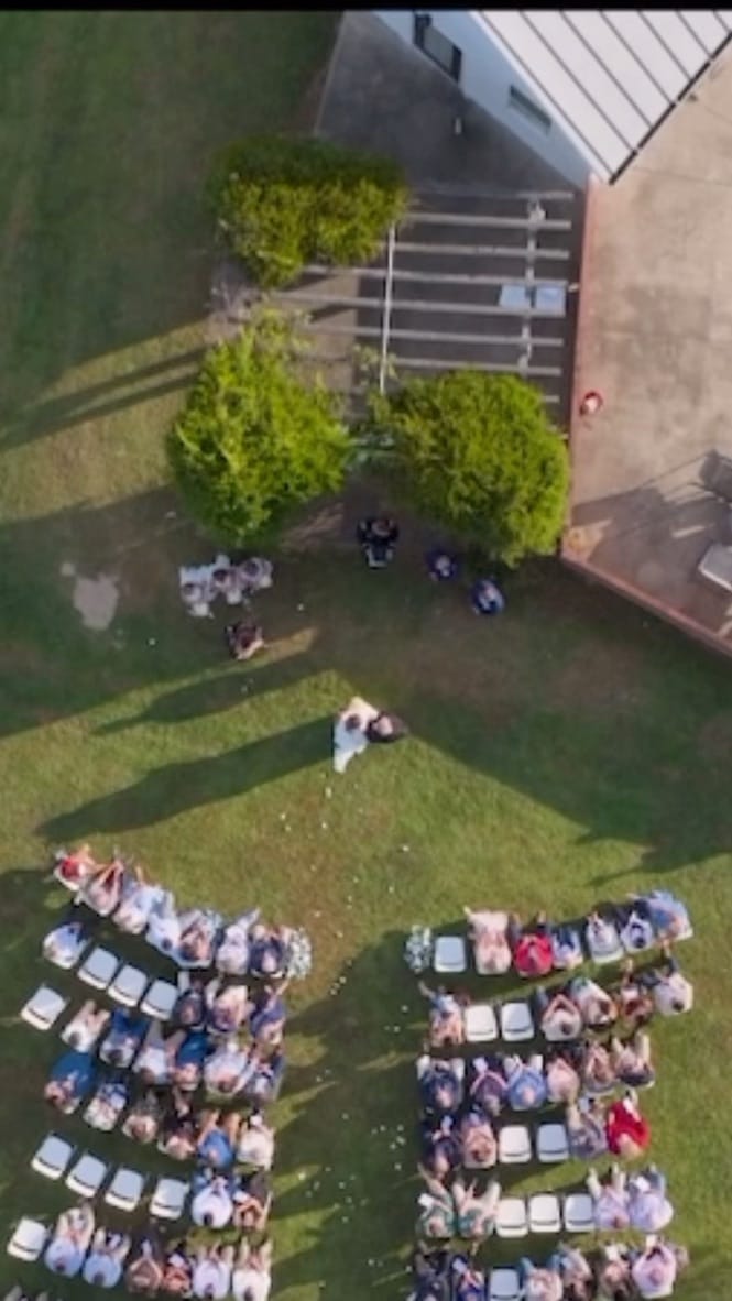 Aerial drone view of an outdoor wedding ceremony at Scarborough House with guests seated in two rows on the green lawn as the couple stands at the aisle surrounded by trees and the historic estate building.