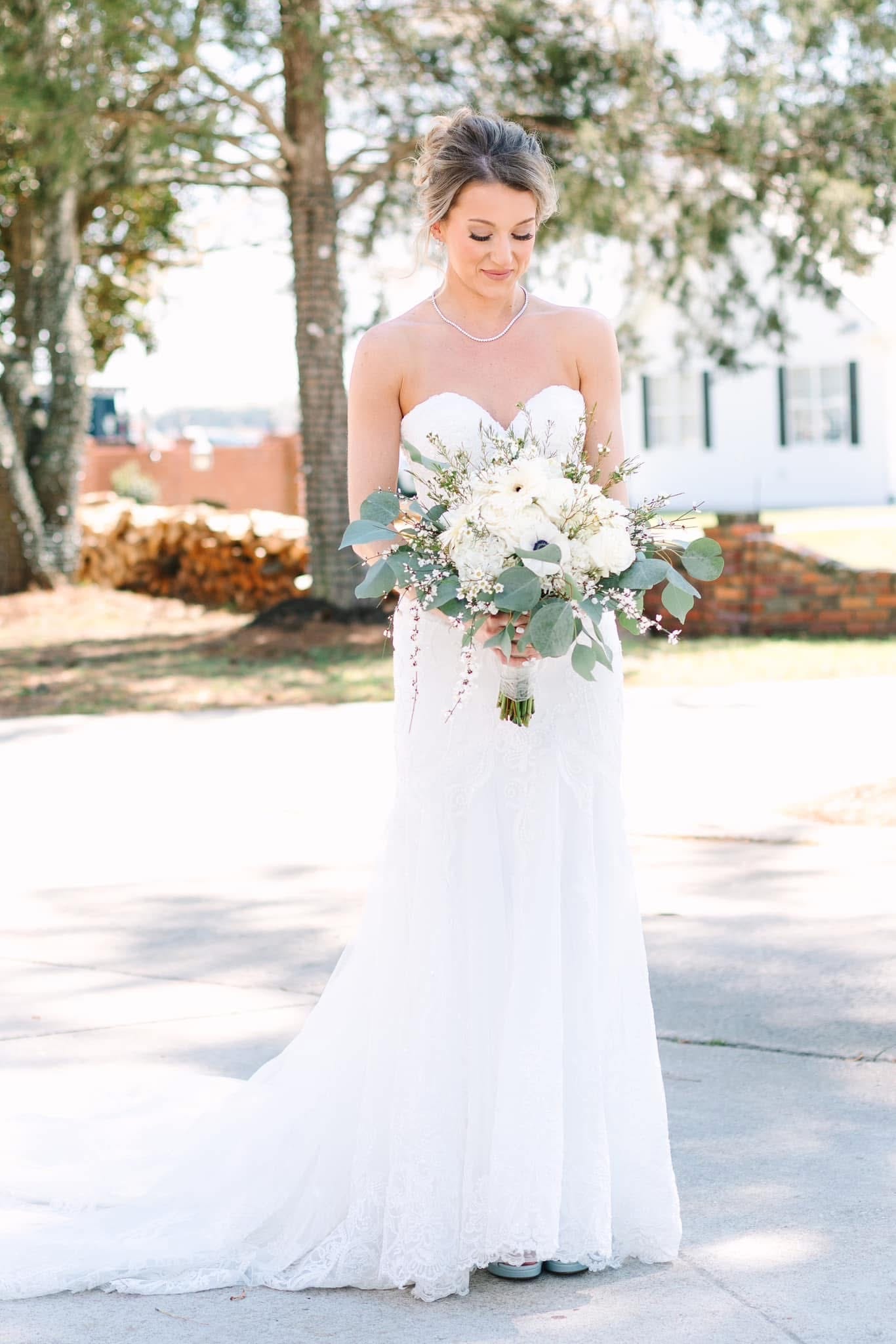 Bride in a strapless lace gown gazes down at her white floral and eucalyptus bouquet while standing beneath oak trees on the grounds of Scarborough House historic estate.