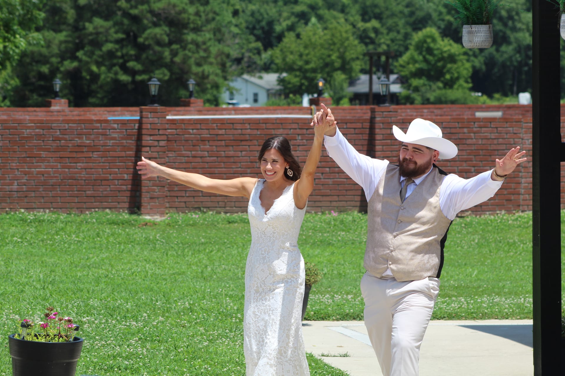 Bride in a white lace gown and groom in a tan vest and white cowboy hat joyfully raise their joined hands as they make their grand entrance on the ceremony lawn at Scarborough House.